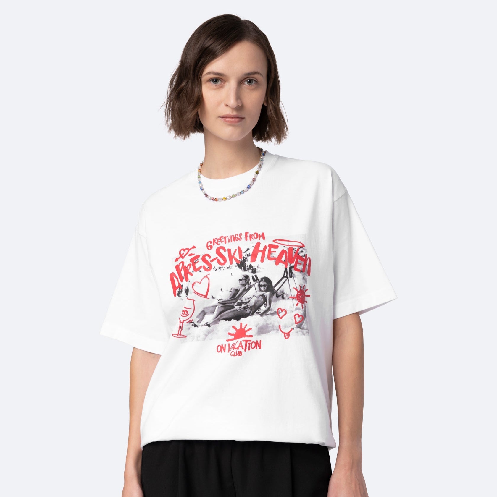 Eine Frau mit kurzen braunen Haaren trägt ein weißes grafisches T-Shirt mit roter und schwarzer Aufschrift "Greetings from Apres-Ski Heaven" und eine schwarze Hose und steht vor einem schlichten hellgrauen Hintergrund.