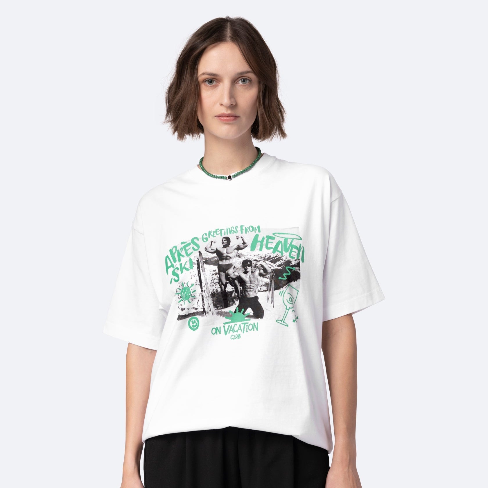 Eine Frau mit kurzen braunen Haaren trägt ein weißes grafisches T-Shirt mit grünem und schwarzem Text und Bildern, auf denen "Greetings From Heaven" und "On Vacation Club" zu lesen sind, gepaart mit einer schwarzen Hose. Sie steht vor einem schlichten hellen Hintergrund.