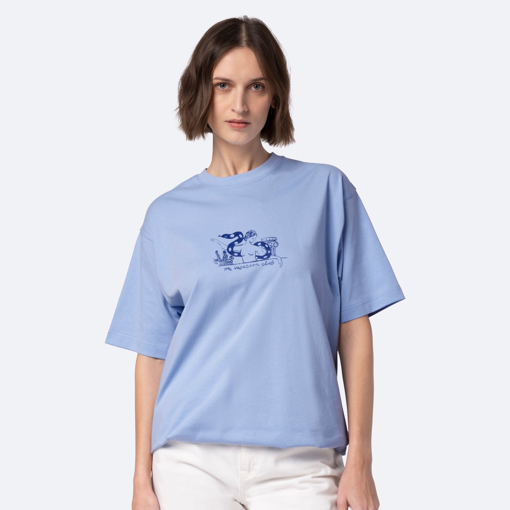 Eine Frau mit kurzen braunen Haaren trägt ein locker sitzendes hellblaues T-Shirt mit einer blauen Grafik und Text, gepaart mit einer weißen Hose, vor einem schlichten hellen Hintergrund.