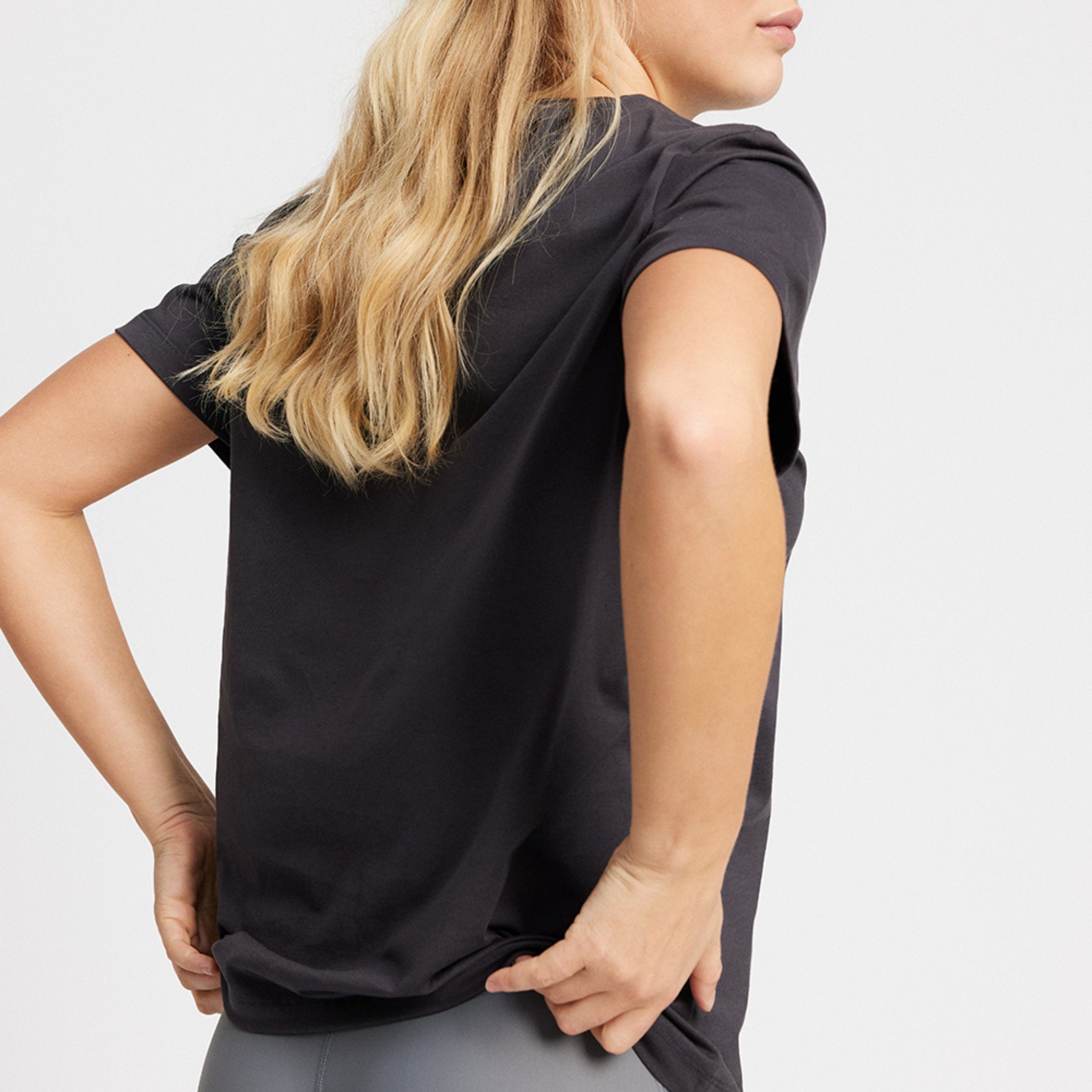 Frau mit langen blonden Haaren trägt ein schwarzes T-Shirt und helle Leggings, seitlich fotografiert.