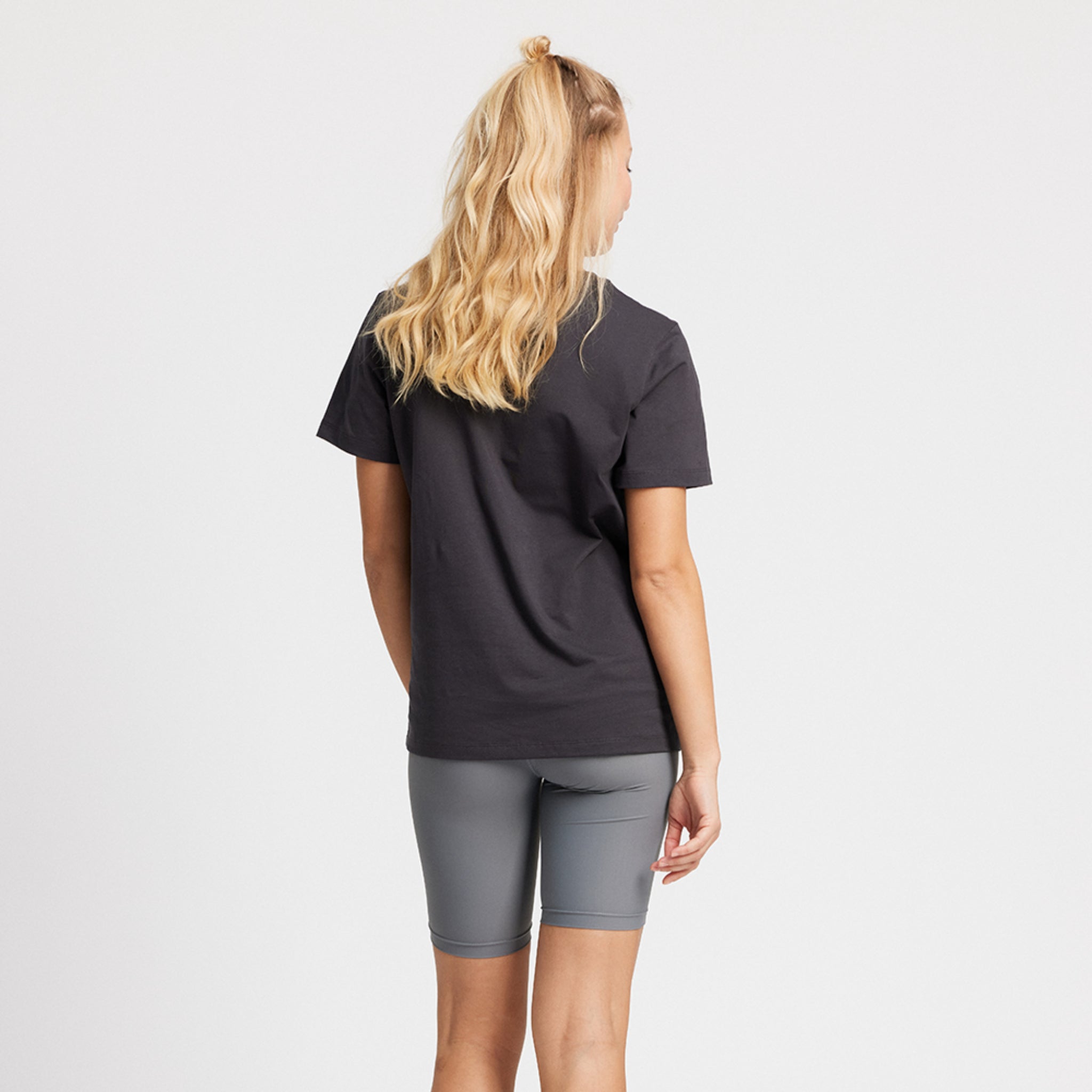 Frau mit langen blonden Haaren trägt ein schwarzes T-Shirt und graue Shorts, steht mit dem Rücken zur Kamera.