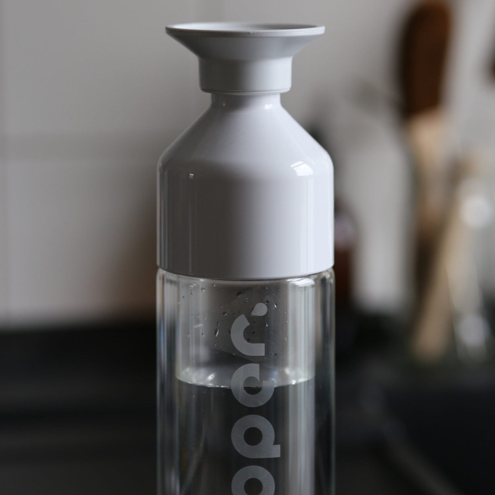 Durchsichtige Flasche mit weißem Deckel, teils gefüllt mit Wasser, in einer unscharfen Küchenumgebung.