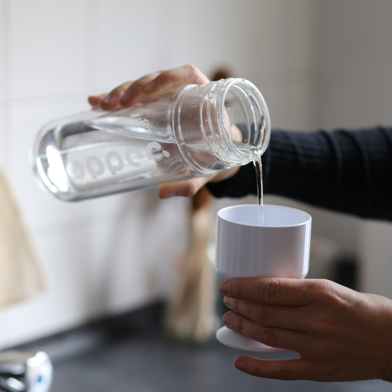 Eine Person gießt Wasser aus einer Glasflasche in einen weißen Becher.