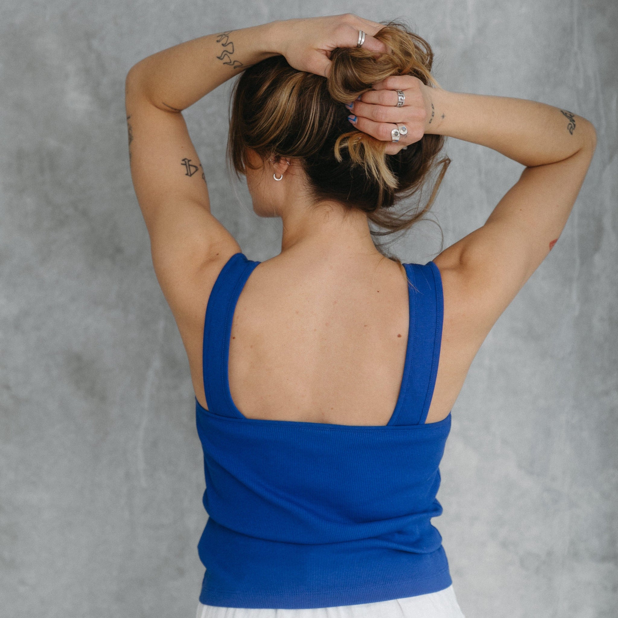 Person mit blauen Tanktop bindet sich die Haare, zeigt Tätowierungen und Schmuck an den Händen, vor grauem Hintergrund.
