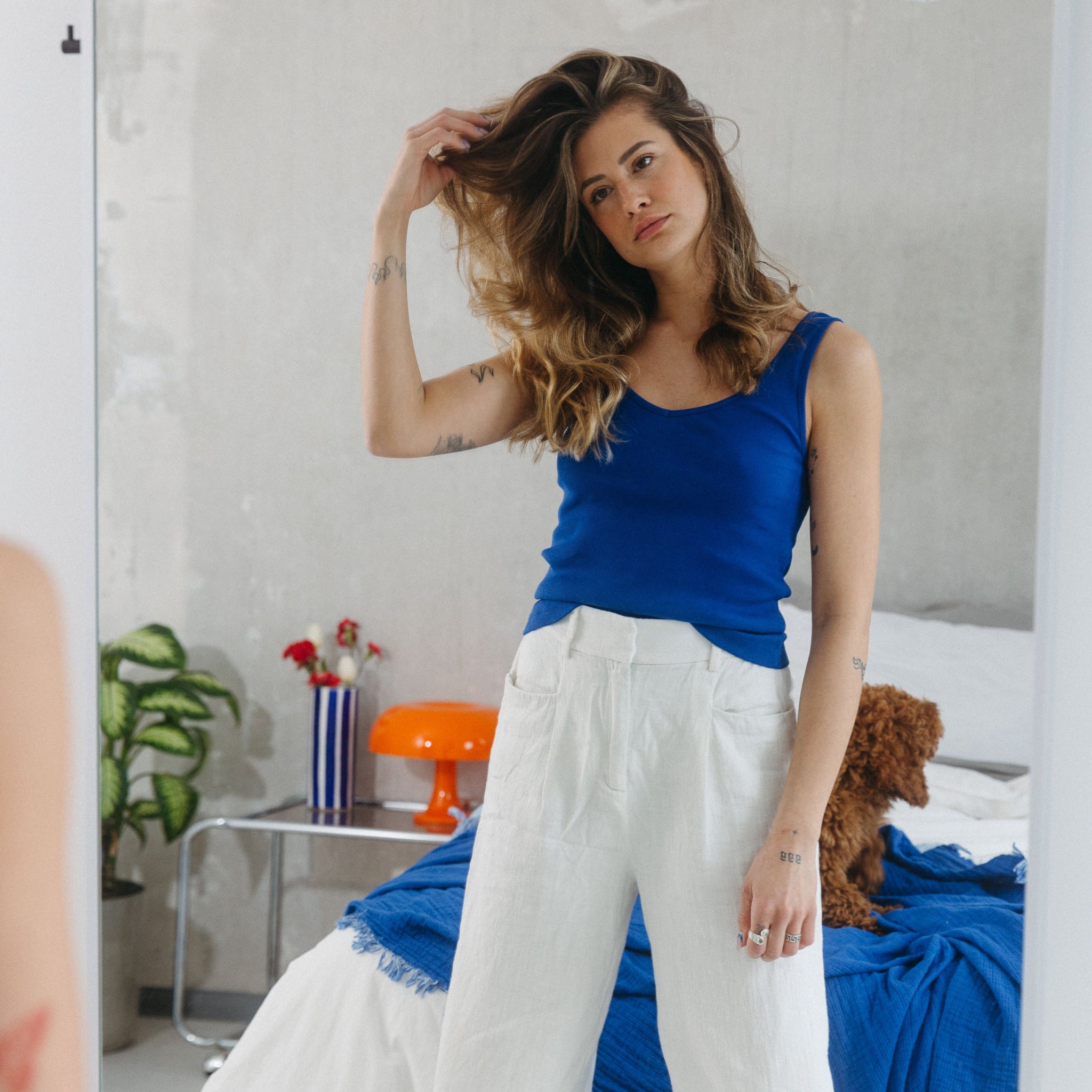 Frau in blauem Top und weißer Hose steht vor einem Spiegel, hebt ihr Haar, Pflanze und roter Lampenschirm im Hintergrund.