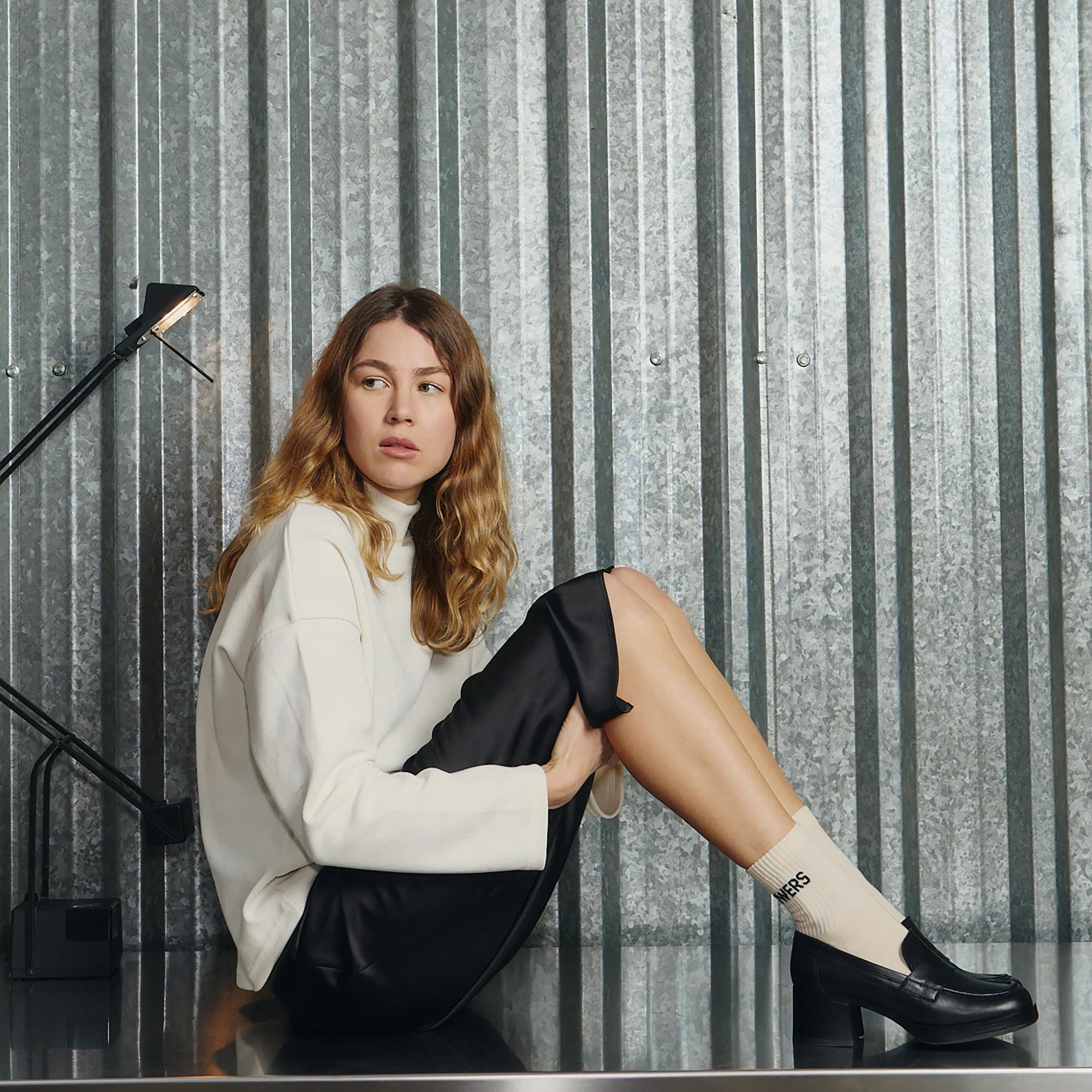 Eine Frau mit gewelltem Haar sitzt auf einer Metallfläche vor einer Wellblechwand und trägt einen weißen Rollkragenpullover, einen schwarzen Rock, schwarze Halbschuhe und weiße Socken. Neben ihr befindet sich eine schwarze Schreibtischlampe.