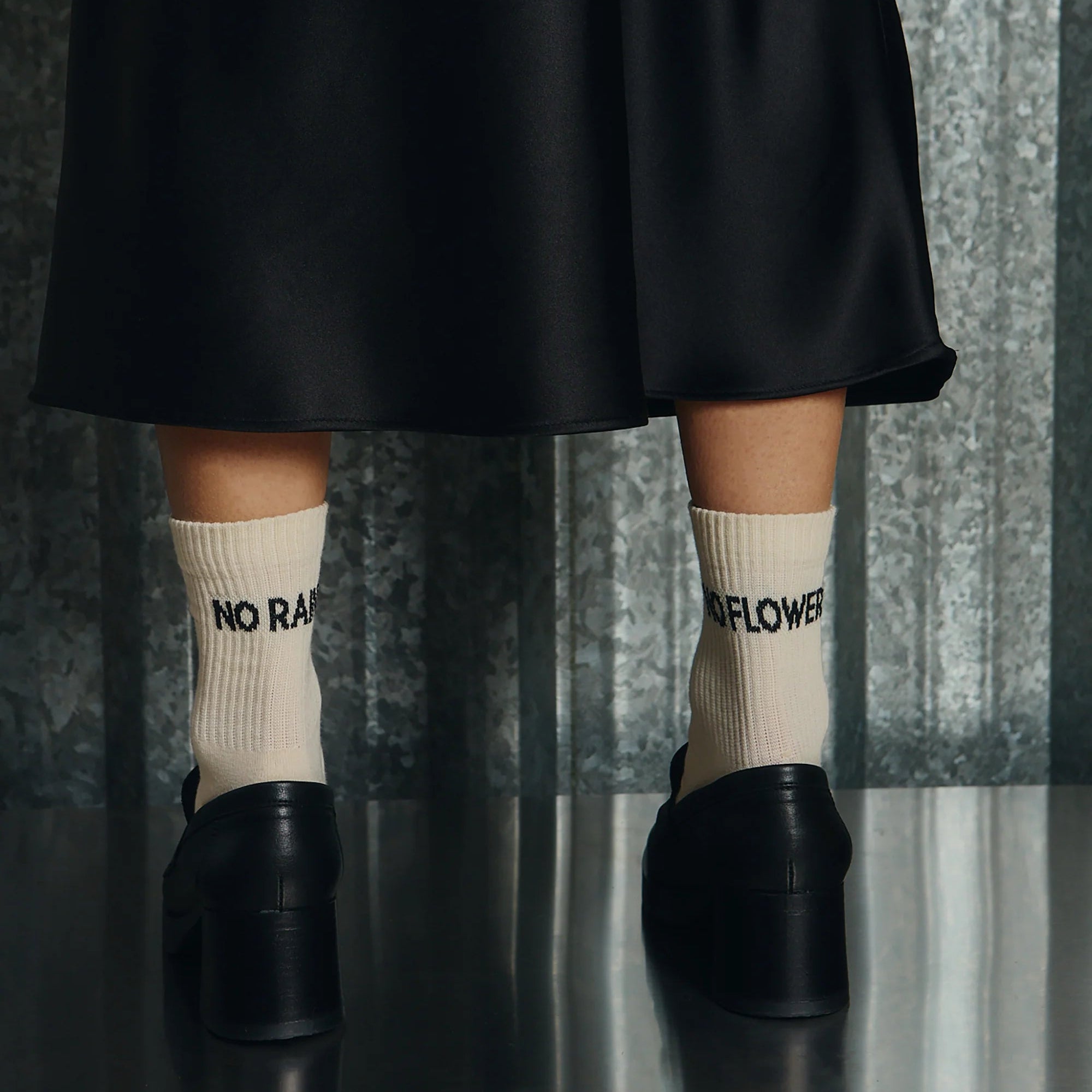 Eine Person, die cremefarbene Socken mit der schwarzen Aufschrift NO RAIN und NO FLOWERS und schwarze Schuhe mit Absätzen trägt, steht auf einem reflektierenden Boden und trägt einen schwarzen Rock vor einem strukturierten metallischen Hintergrund.