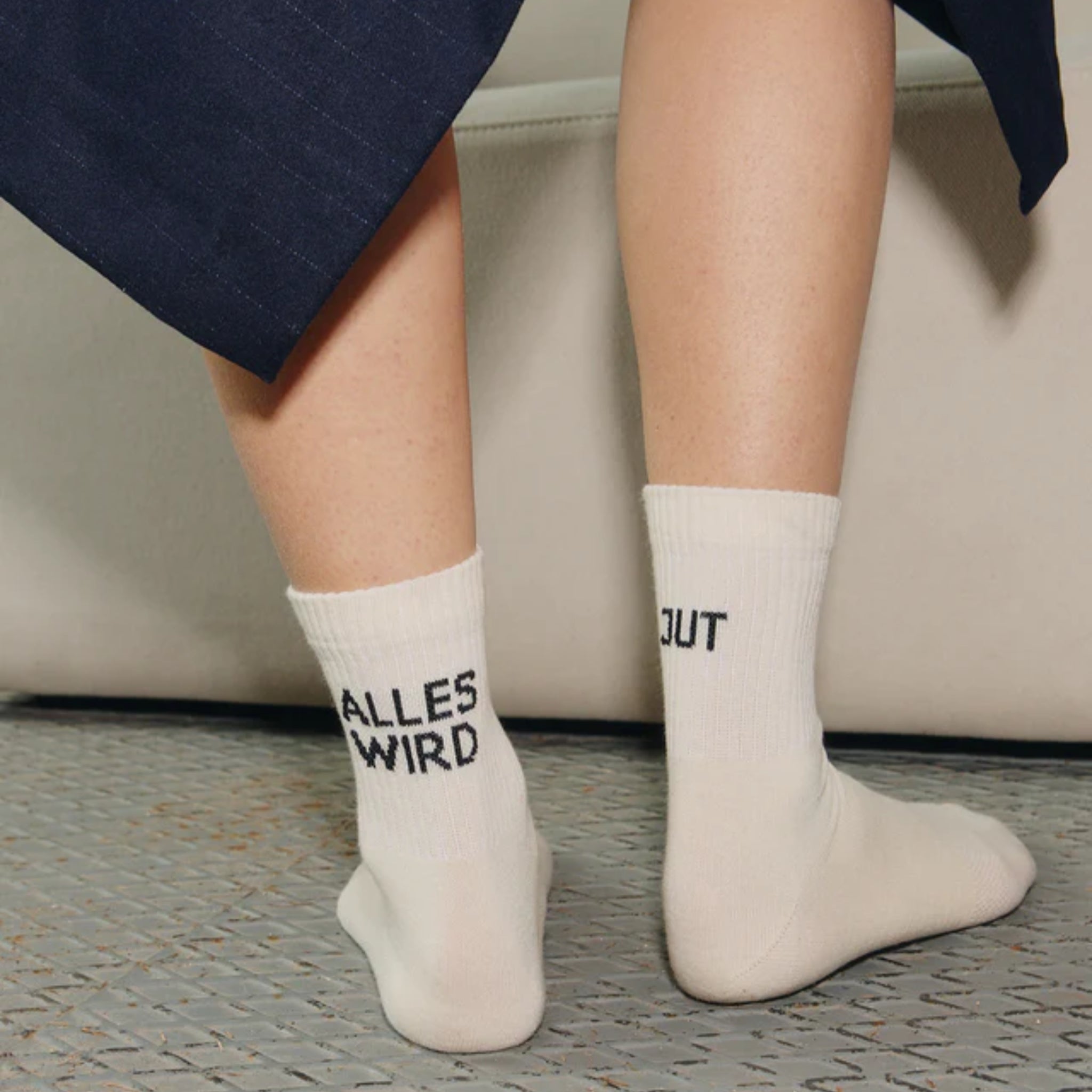 Eine Person, die einen dunklen Rock und weiße Socken mit der schwarzen Aufschrift "ALLES WIRD" auf einer Socke und "GUT" auf der anderen trägt, steht auf einer strukturierten Fläche.
