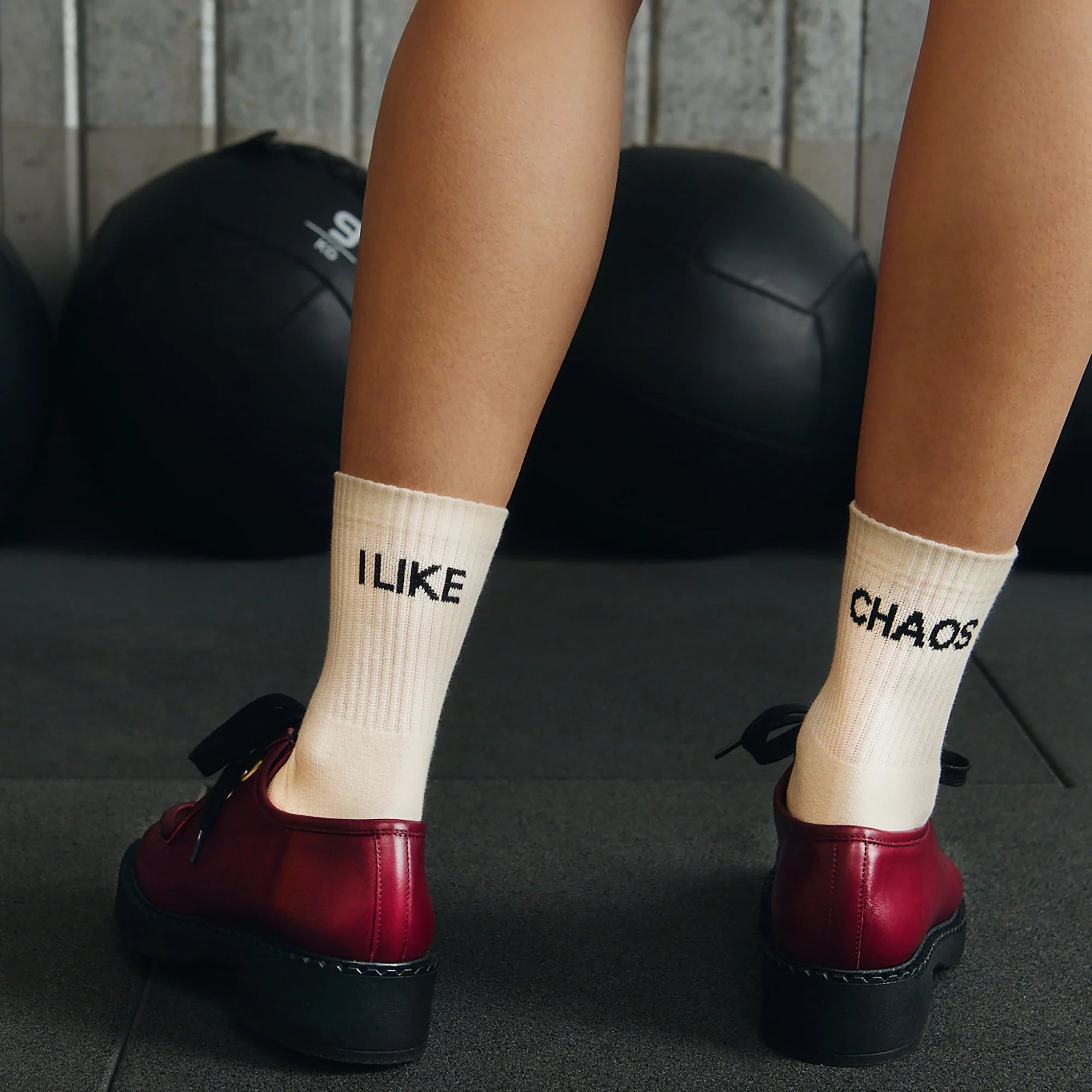 Eine Person, die cremefarbene Socken mit der Aufschrift I LIKE CHAOS und rote Schuhe trägt, steht auf dem Boden einer Turnhalle mit schwarzen Gymnastikbällen im Hintergrund.