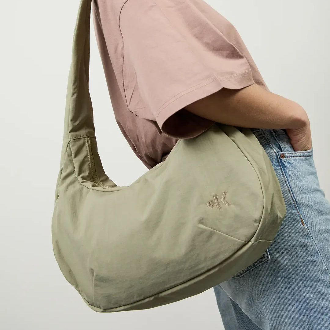 Eine Person mit roten Locken trägt ein lockeres lilafarbenes T-Shirt und hellblaue Jeans und hat eine große, hellolivgrüne Umhängetasche dabei. Der Hintergrund ist einfarbig und hell.