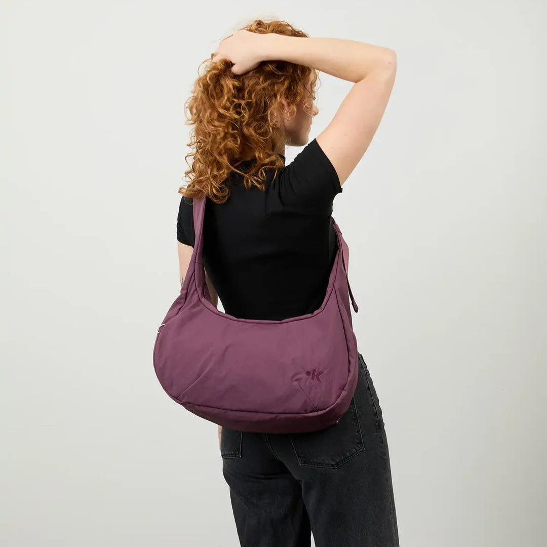 Eine Person mit lockigem rotem Haar, die ein schwarzes Hemd und dunkle Jeans trägt, steht mit dem Rücken zur Kamera. Sie hat einen Arm erhoben und trägt eine große lila Umhängetasche. Der Hintergrund ist einfarbig und hell.