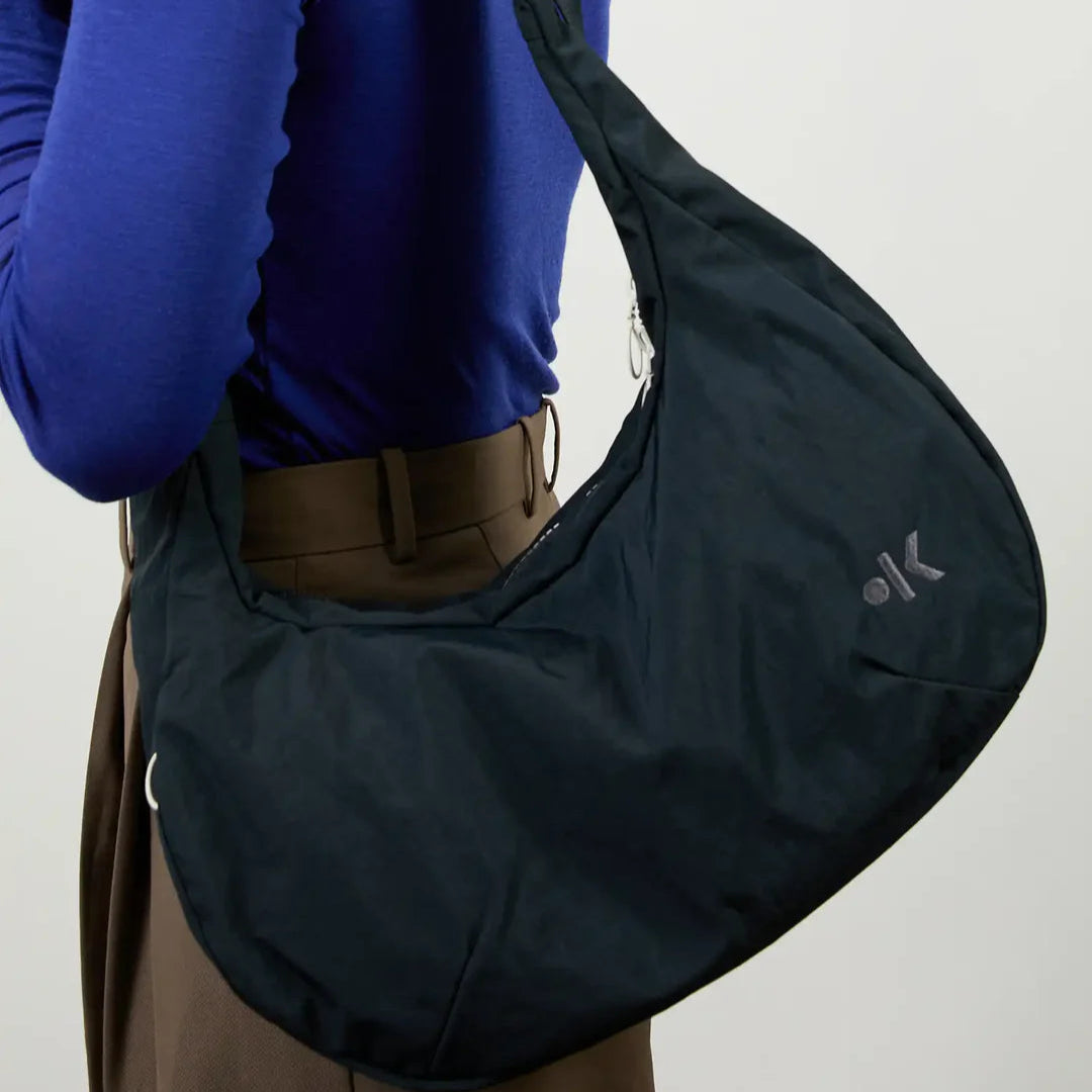 Eine Person mit lockigem Haar, bekleidet mit einem langärmeligen blauen Oberteil und einer braunen Hose, trägt eine große schwarze Umhängetasche mit einem kleinen weißen Logo an der Seite.