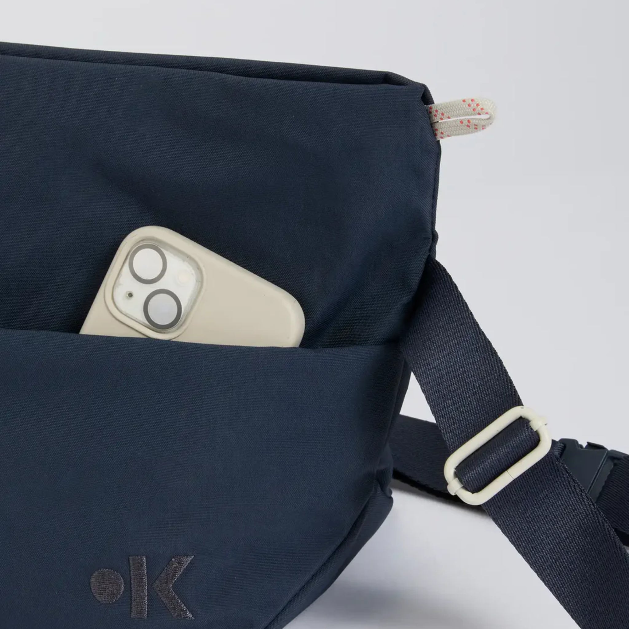 Ein cremefarbenes Smartphone, das teilweise in die Vordertasche einer dunkelnavyfarbenen Stofftasche mit einem verstellbaren Riemen und einer kleinen gemusterten Schlaufe an der oberen Ecke gesteckt wurde.