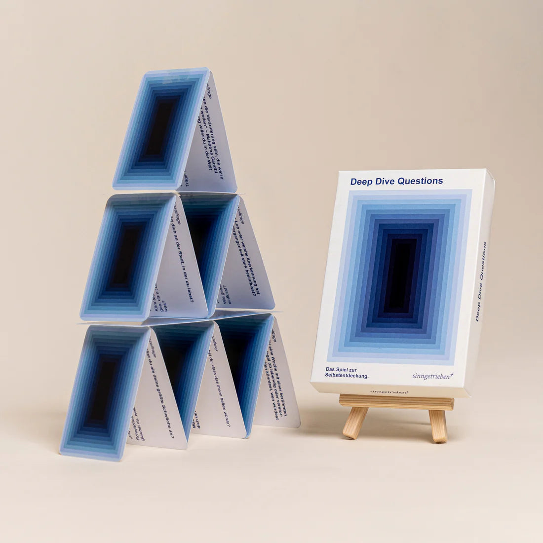 Ein Stapel Karten in einer Pyramide neben einer Schachtel mit der Aufschrift Deep Dive Questions auf einer kleinen hölzernen Staffelei, beide mit einem blauen geometrischen Muster auf beigem Hintergrund.