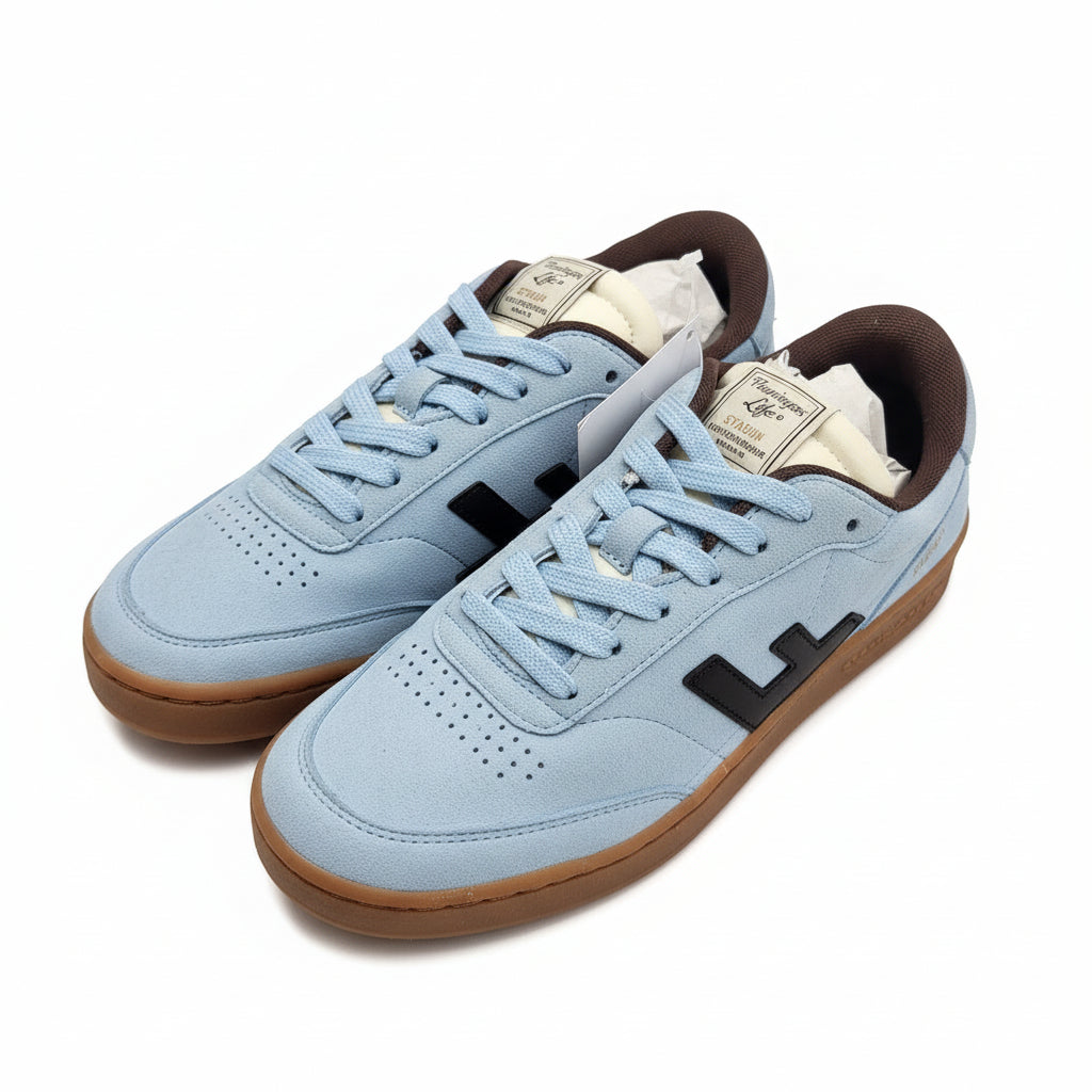 Ein Paar hellblaue Turnschuhe mit braunen Sohlen und schwarzen Akzenten stehen auf einem grauen, leicht strukturierten Boden. Die Schuhe haben eine braune Innenausstattung und Etiketten an den Schnürsenkeln.