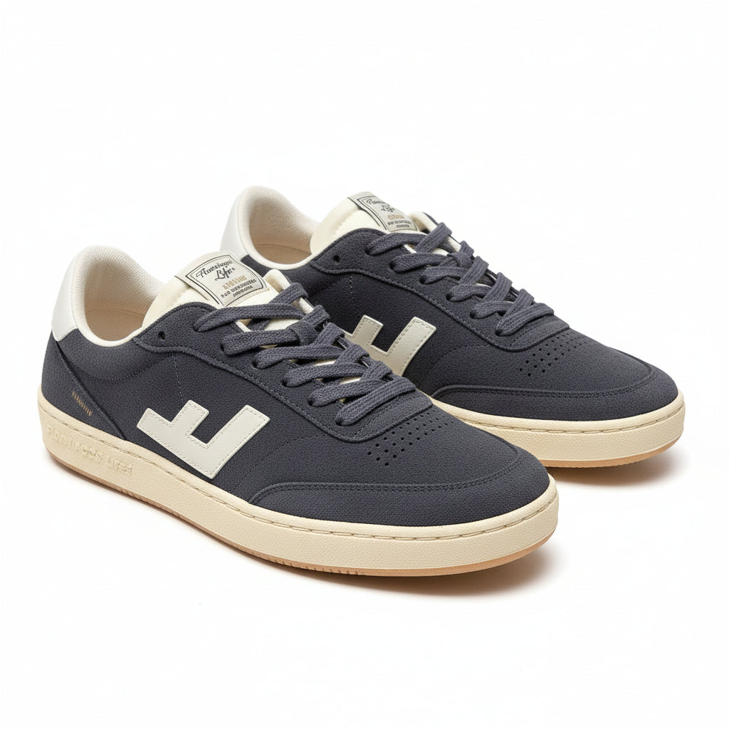 Ein Paar dunkelgraue und weiße Turnschuhe mit cremefarbenen Sohlen stehen auf einem grauen Boden. Ein Schuh ist mit weißem Papier ausgestopft, und beide haben ein Etikett auf der Zunge, auf dem Maison Mihara Yasuhiro steht.