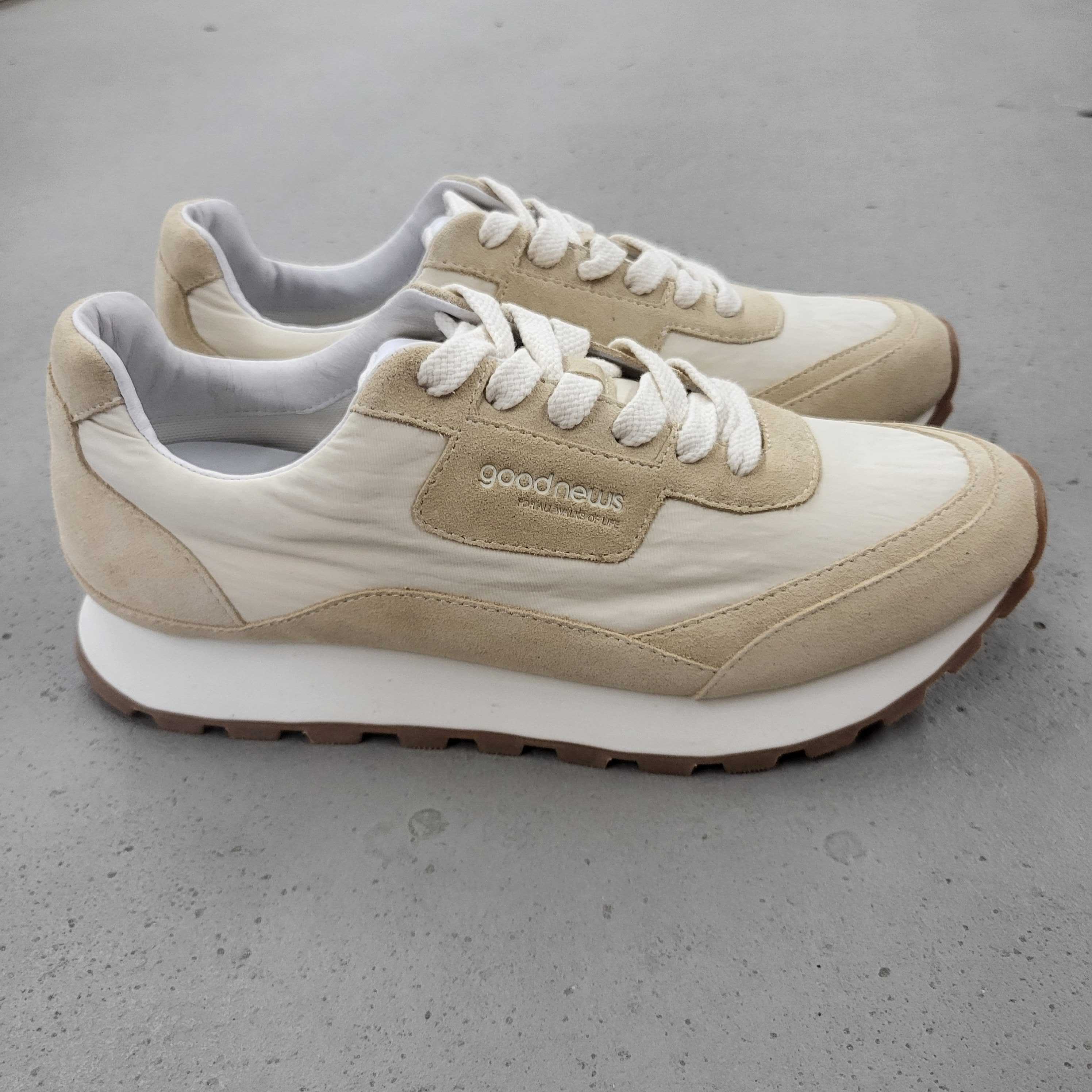 Ein Paar beige-weiße Turnschuhe mit weißen Schnürsenkeln, braunen Gummisohlen und Goodnews-Branding an der Seite, die auf einer hellgrauen Betonfläche stehen.