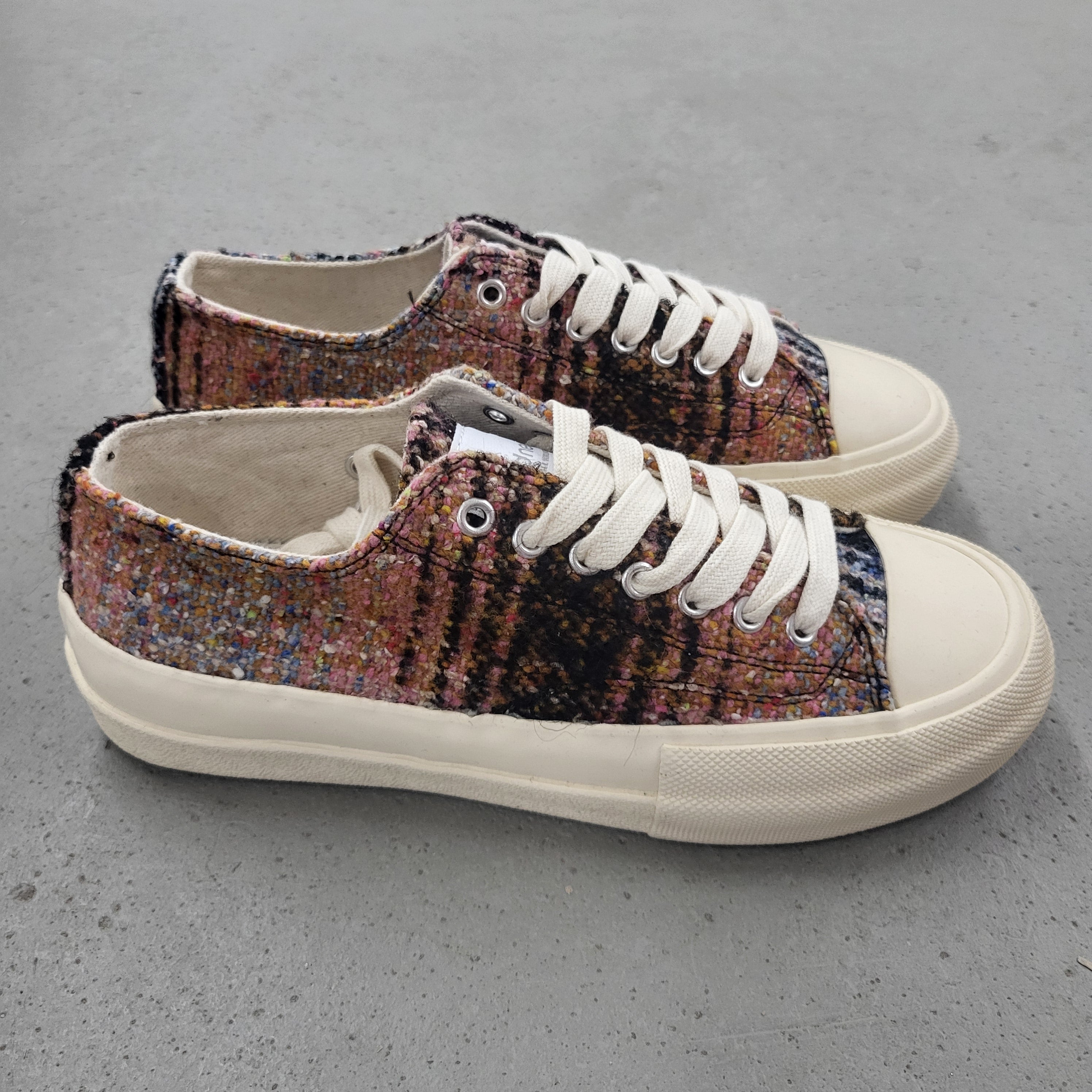 Ein Paar Low-Top-Sneaker mit weißer Sohle und Schnürsenkeln, deren Obermaterial aus buntem, strukturiertem Stoff in Rosa-, Schwarz-, Blau- und Beigetönen besteht und die auf einer grauen Betonfläche stehen.