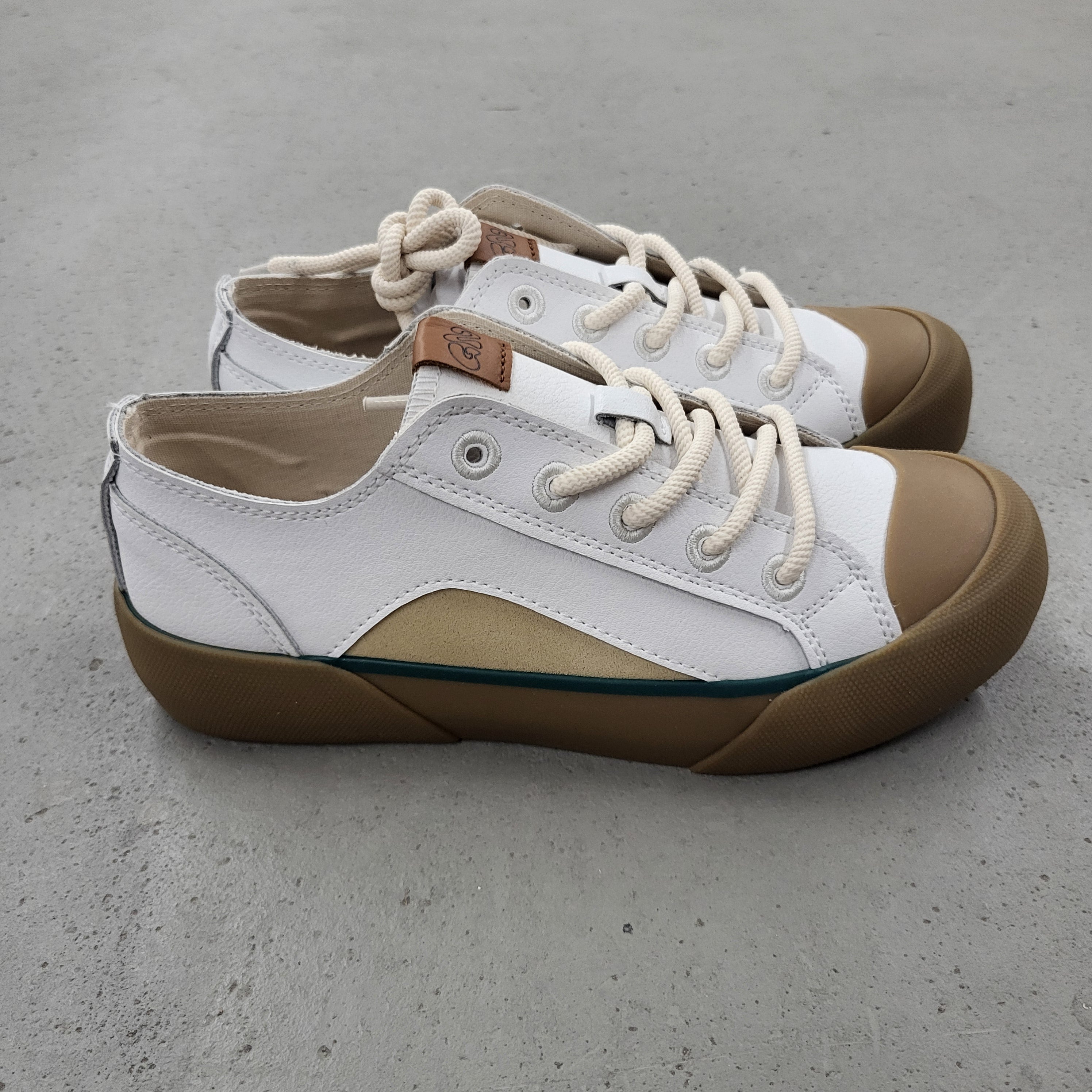 Ein Paar weißer Segeltuch-Sneakers mit hellbraunen Gummikappen und -sohlen, beigen Schnürsenkeln und einem kleinen braunen Lederflicken in der Nähe des Knöchels, die auf einer grauen Betonfläche stehen.