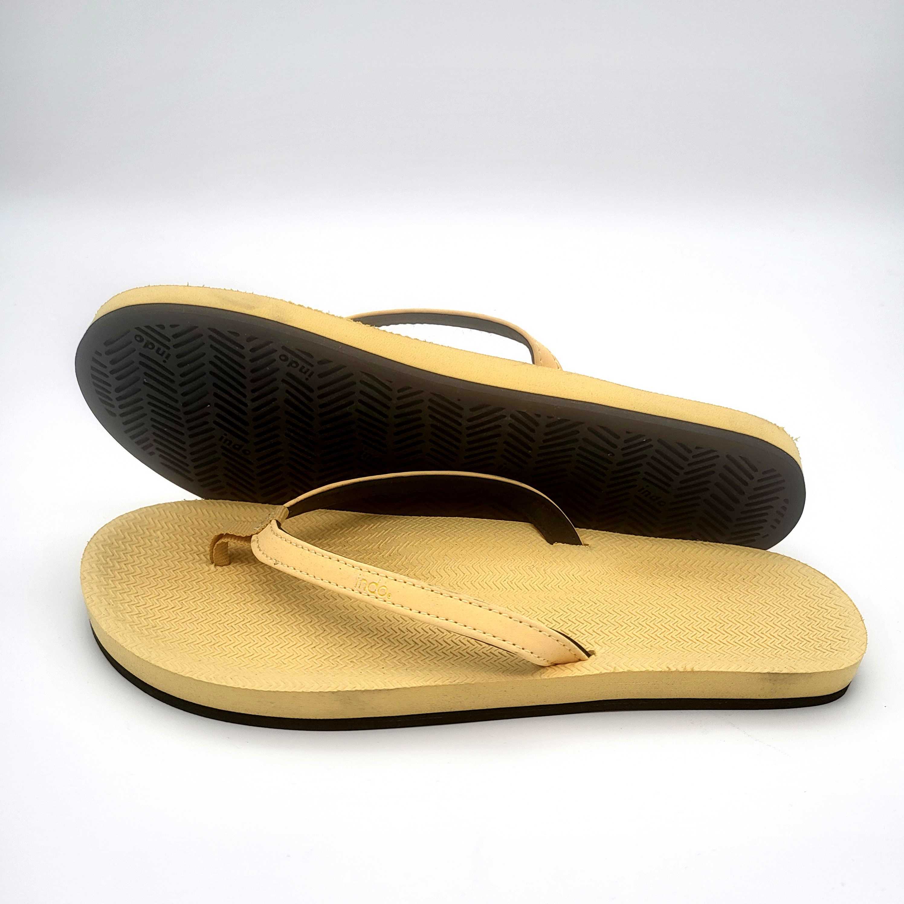 Ein Paar beigefarbene Flip-Flops mit schwarzer Sohle; eine Sandale liegt flach, während die andere auf den Rand gestützt ist, so dass der strukturierte Boden zu sehen ist. Der Hintergrund ist einfarbig weiß.