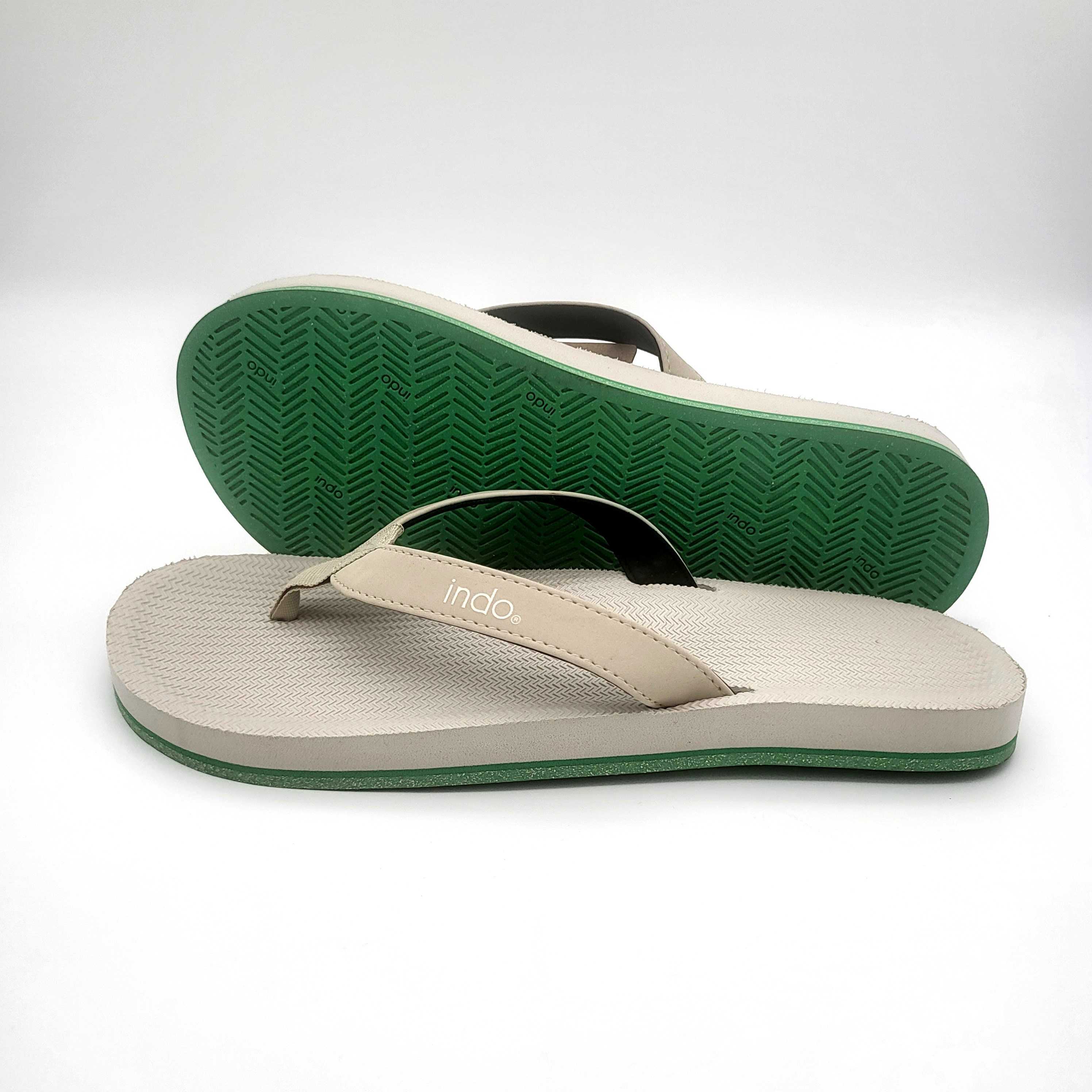 Ein Paar beigefarbene Flip-Flops mit grünen Sohlen. Eine Sandale liegt flach, die andere ist dagegen gestützt, so dass die strukturierte grüne Sohle zu sehen ist.