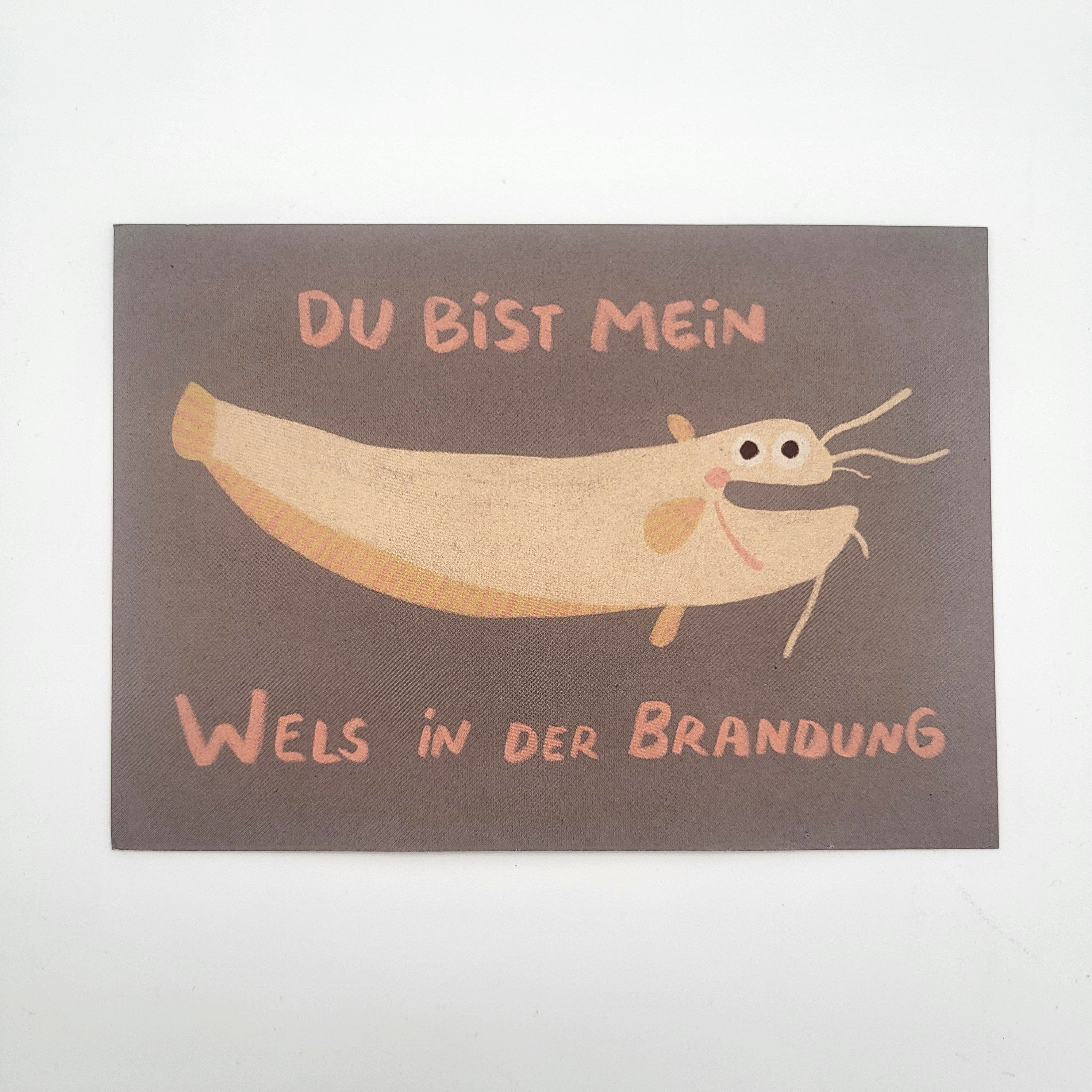 Auf einer grauen Karte ist ein Cartoon-Wels mit einem lächelnden Gesicht abgebildet. Darüber und darunter steht ein deutscher Text: DU BIST MEIN WELS IN DER BRANDUNG, in pfirsichfarbenen Buchstaben.