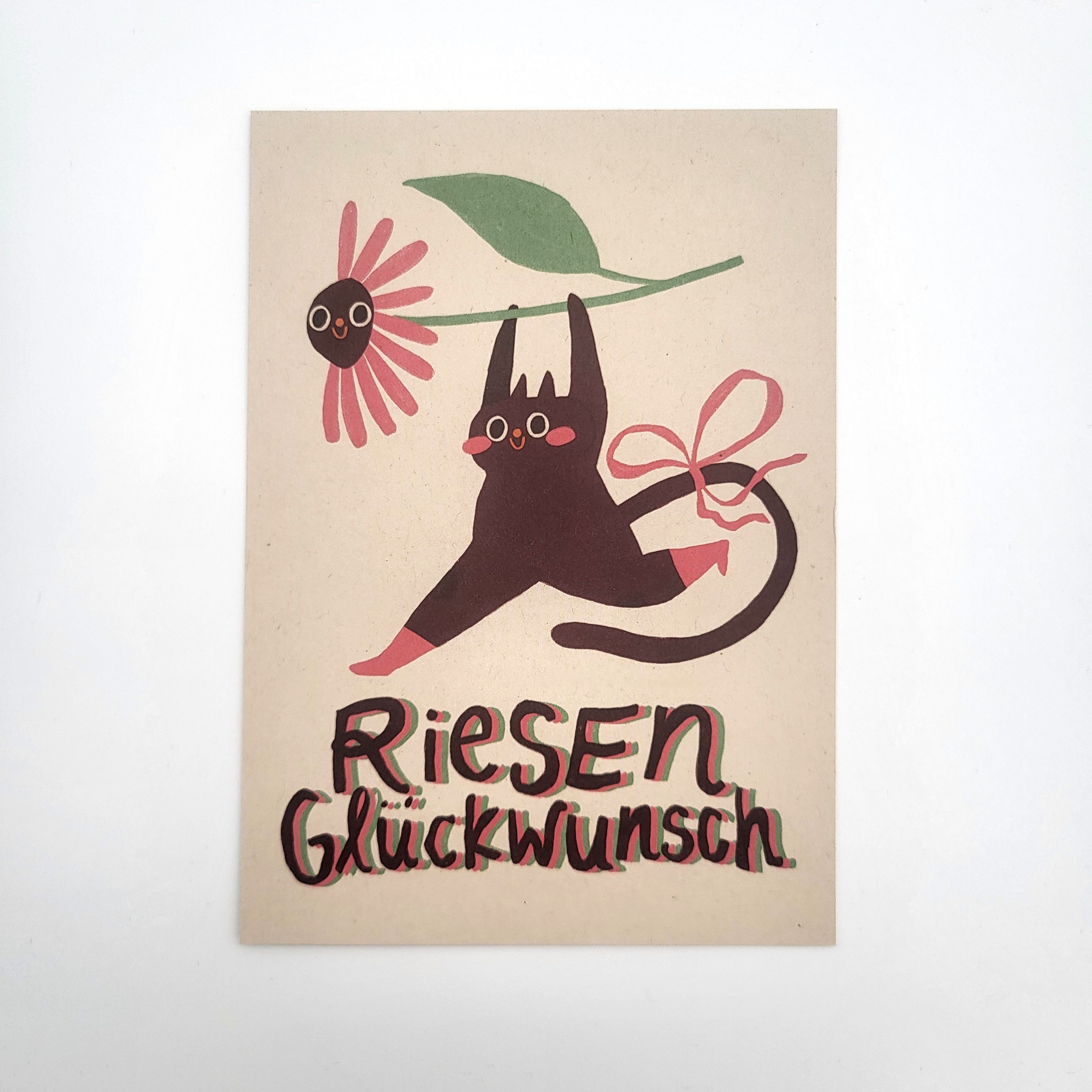 Eine verspielte Illustration einer schwarzen Katze, die mit ihrem Maul eine rosa Blume hält und am Schwanz ein rosa Band trägt. Unter der Katze stehen in bunten Buchstaben die deutschen Worte Riesen Glückwunsch.