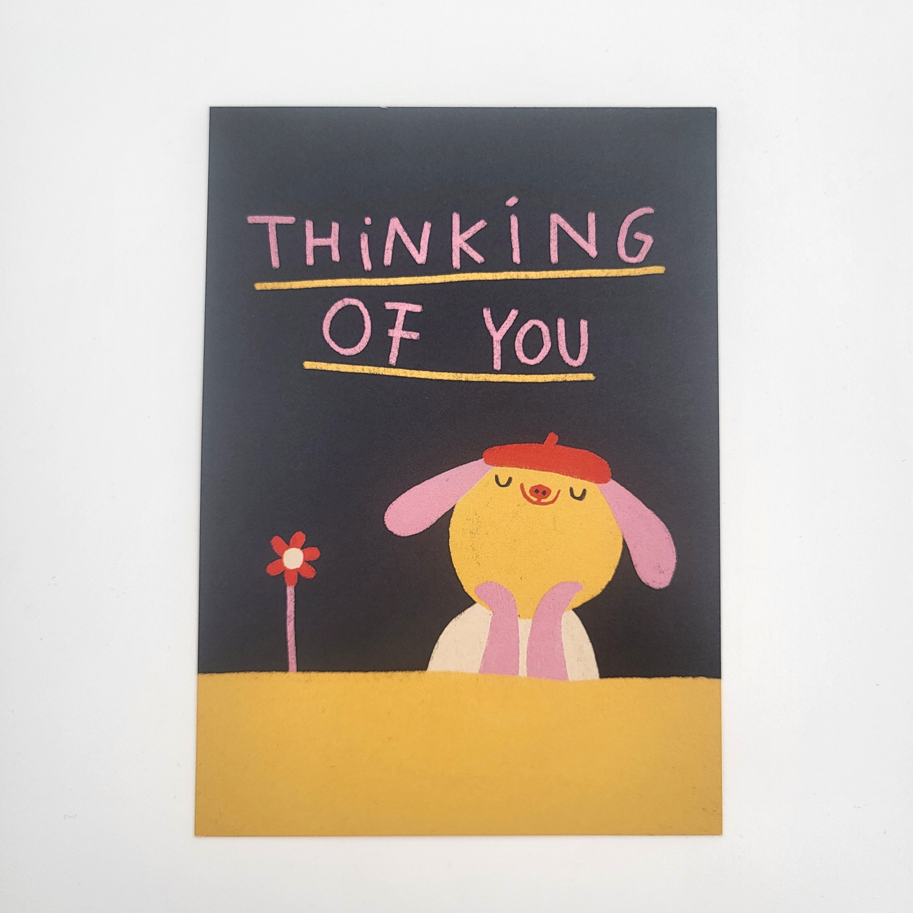 Eine niedliche Zeichnung eines gelben Hundes mit einer roten Baskenmütze und geschlossenen Augen, der neben einer roten Blume sitzt. Darüber steht handgeschriebener Text: THINKING OF YOU auf dunklem Hintergrund.