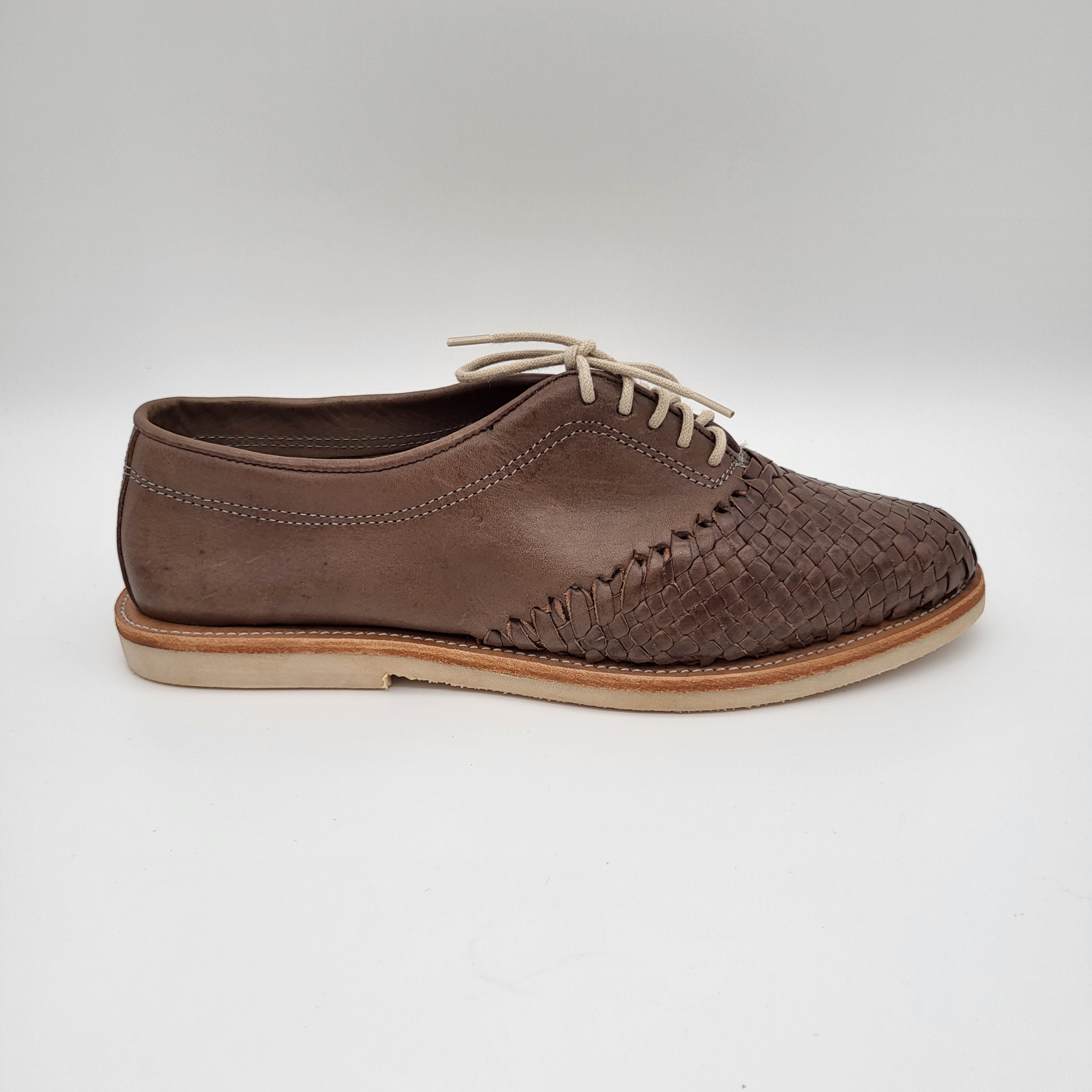 Ein brauner Lederschuh mit einem gewebten Muster auf der Vorderseite, weißen Schnürsenkeln, weißen Nähten und einer hellen Sohle, dargestellt vor einem einfarbigen weißen Hintergrund.