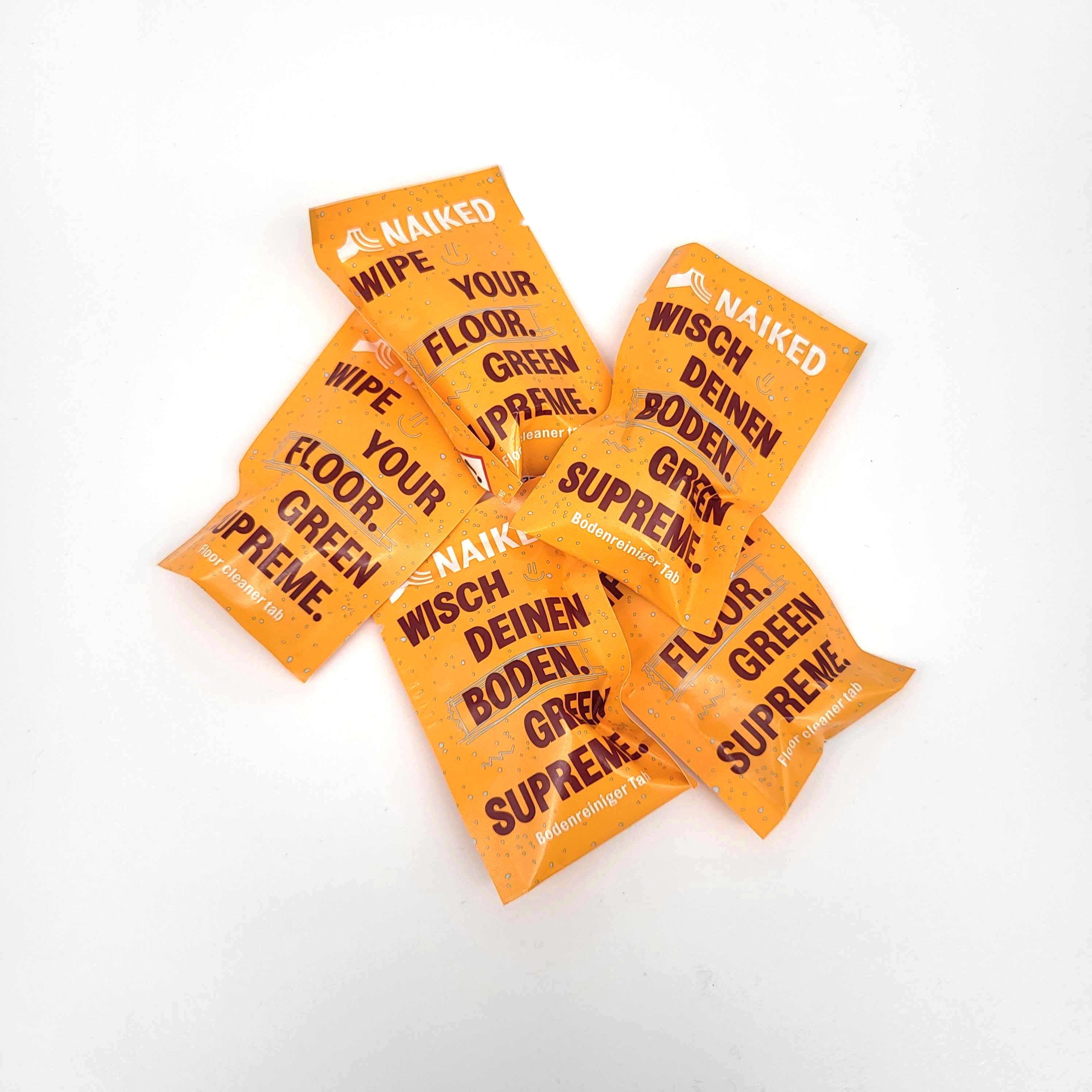 Mehrere orangefarbene Reinigungstücher mit fetter schwarzer und weißer Schrift: NAIKED YOUR WIPE. IHR BODEN. GRÜN SUPREME. Sie liegen verstreut auf einer weißen Fläche.