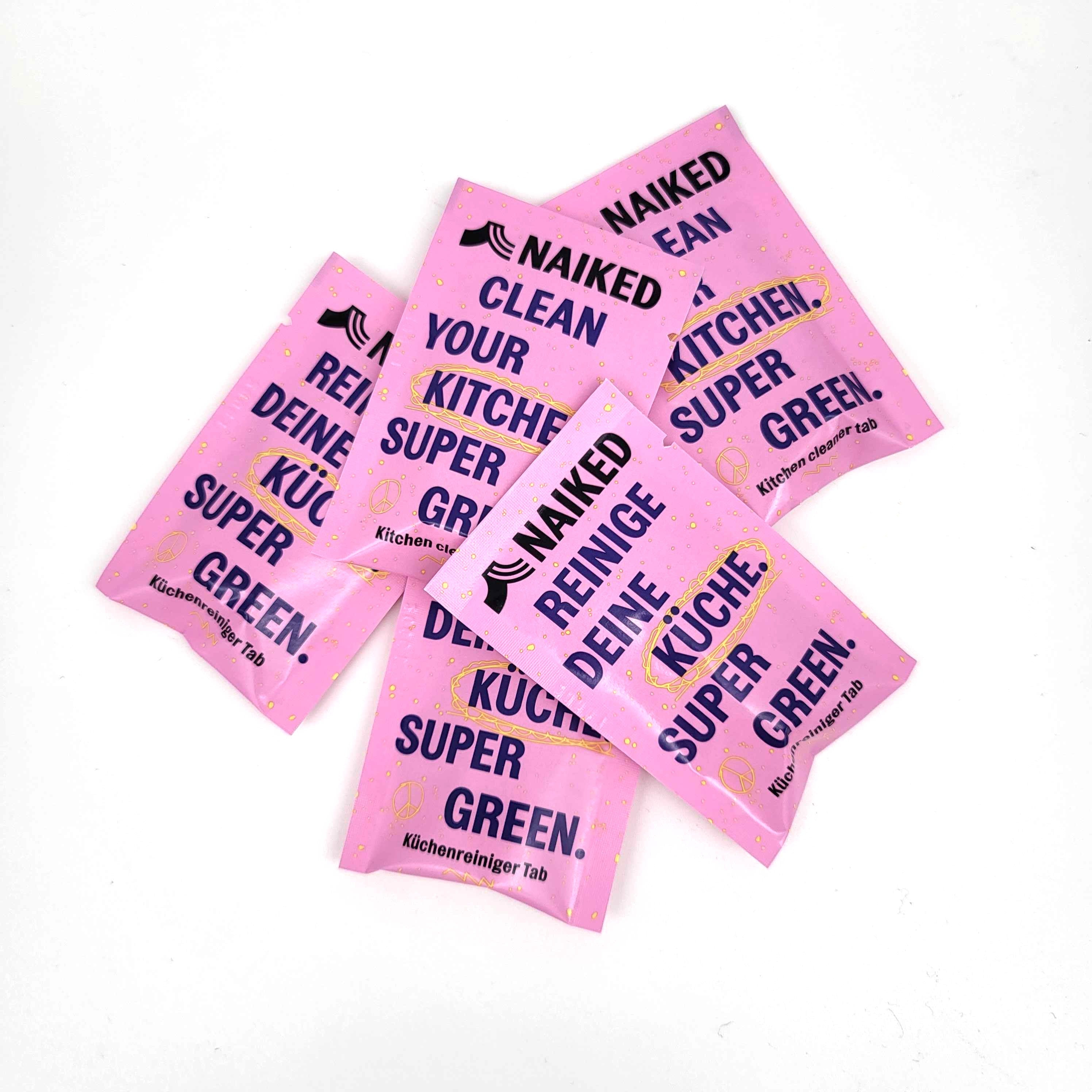 Fünf rosafarbene Küchenreiniger-Tabs mit fettgedruckter schwarzer und gelber Schrift, auf denen NAKED CLEAN YOUR KITCHEN SUPER GREEN! und REINIGE DEINE KÜCHE SUPER GREEN! steht, angeordnet auf einer weißen Fläche.