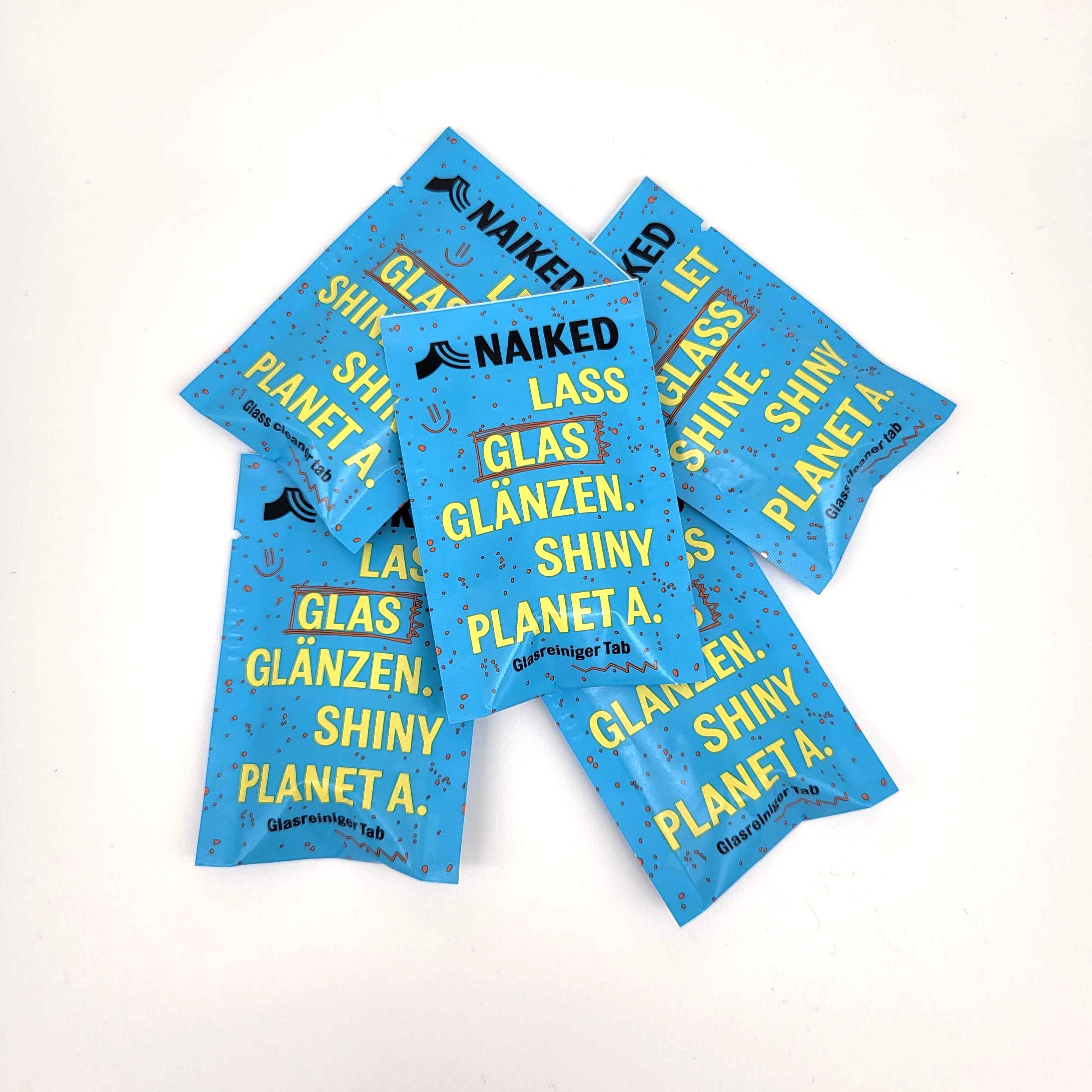 Fünf blaue Päckchen mit der Aufschrift NAIKED und LASS GLAS GLÄNZEN. SHINY PLANET A. sind auf einer weißen Fläche verstreut. Die Päckchen scheinen Glasreinigungstabletten zu sein.