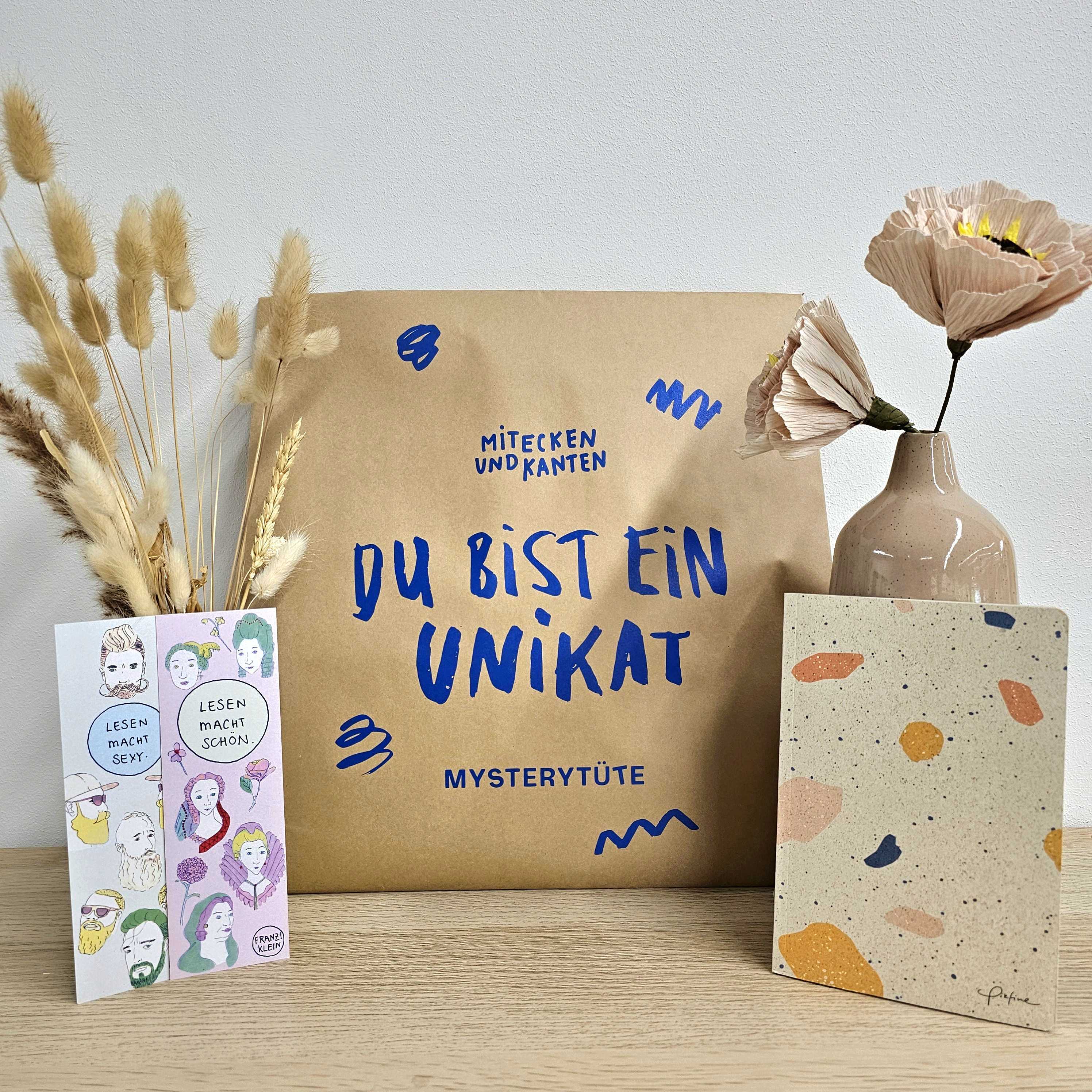Eine braune Papiertüte mit blauem Text, eine beigefarbene Vase mit Trockenblumen und einer rosafarbenen Papierblume, ein illustriertes Büchlein und ein gesprenkeltes beigefarbenes Notizbuch liegen auf einer hellen Holzfläche vor einer schlichten Wand.