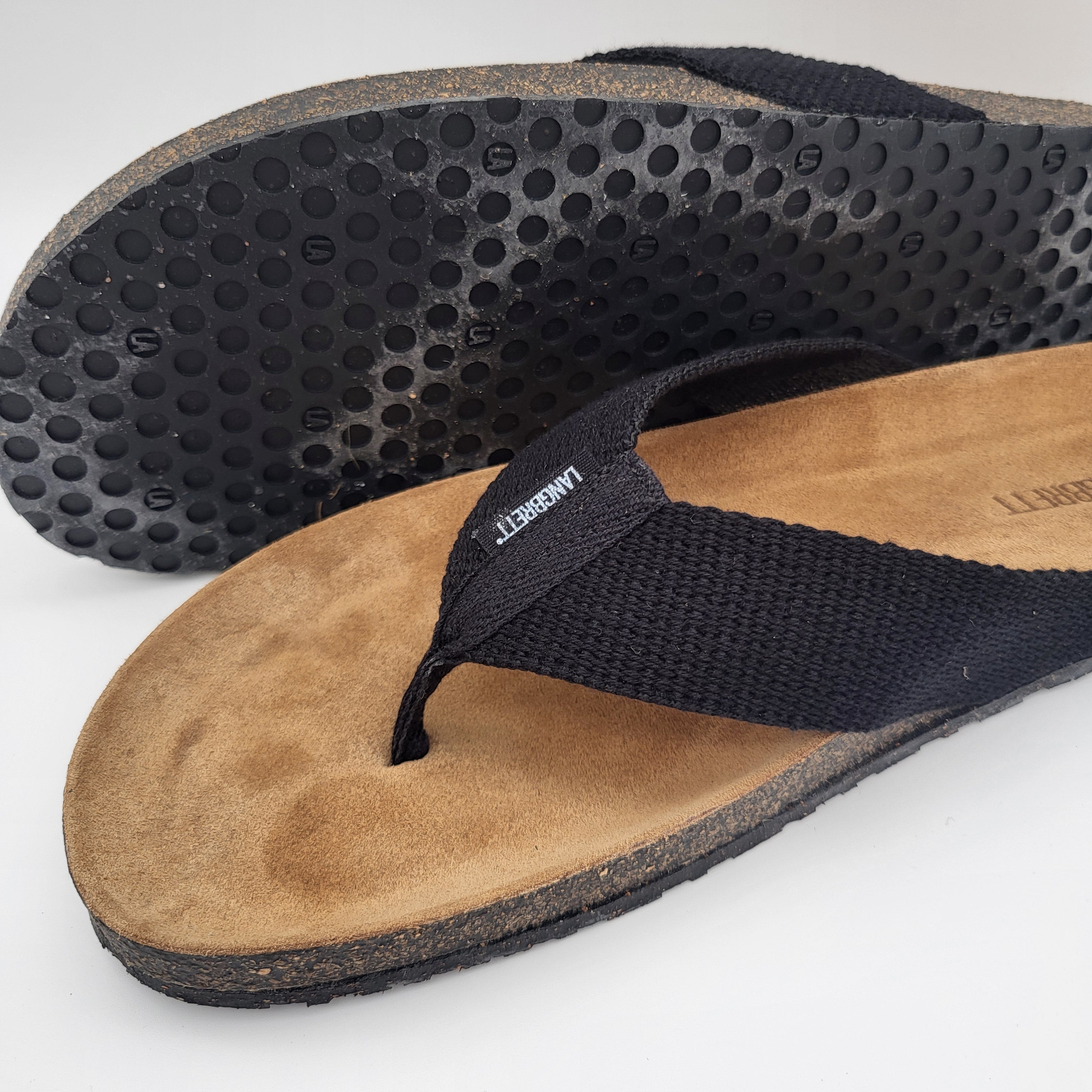 Nahaufnahme zweier schwarzer Flip-Flop-Sandalen mit strukturierter Gummisohle und hellbraunem Wildlederfußbett, mit Stoffriemen und sichtbarem Markenetikett.