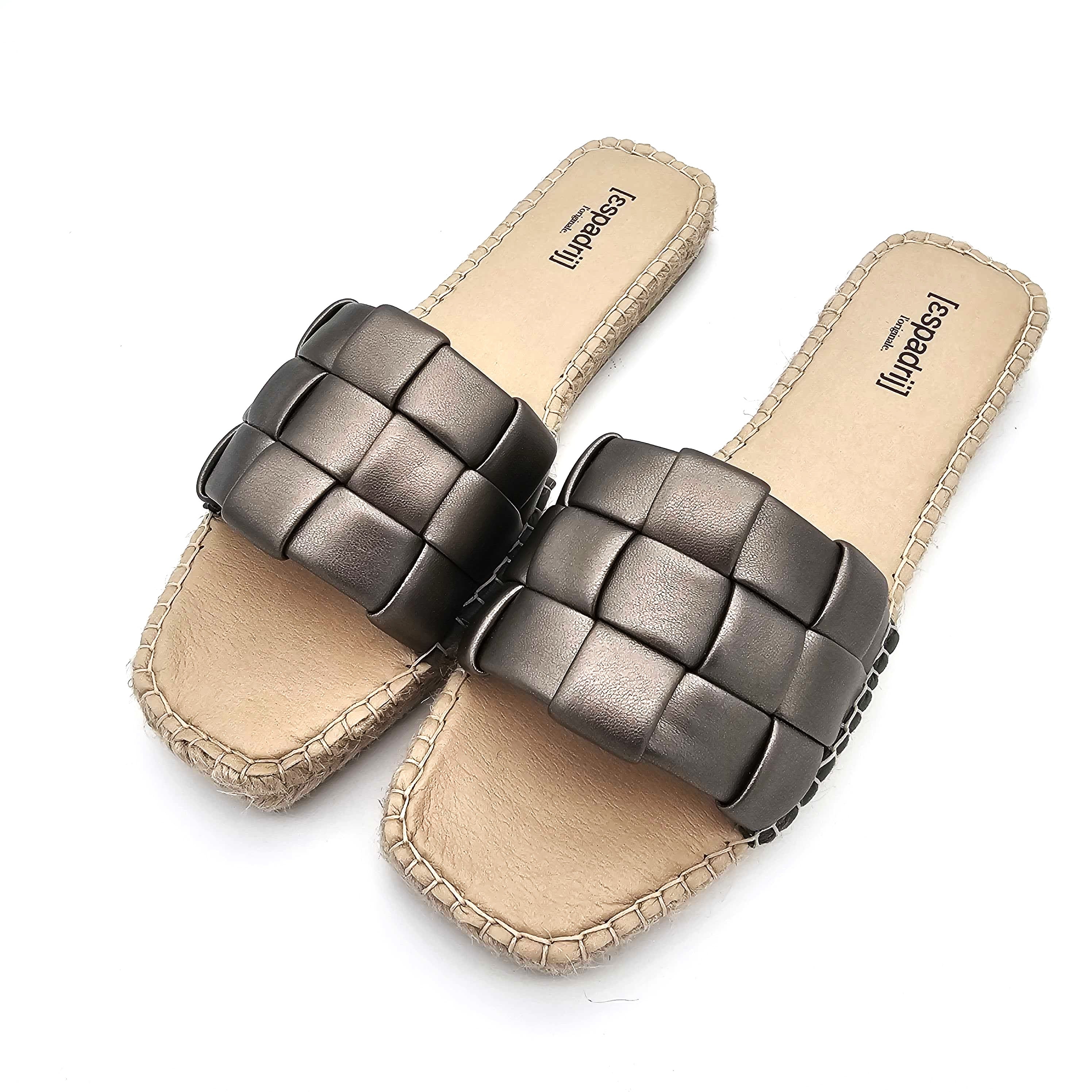 Ein Paar flache Damensandalen mit hellbraunen Sohlen und breiten, gewebten Metallriemen; die Nähte sind entlang der Sohlenränder sichtbar.