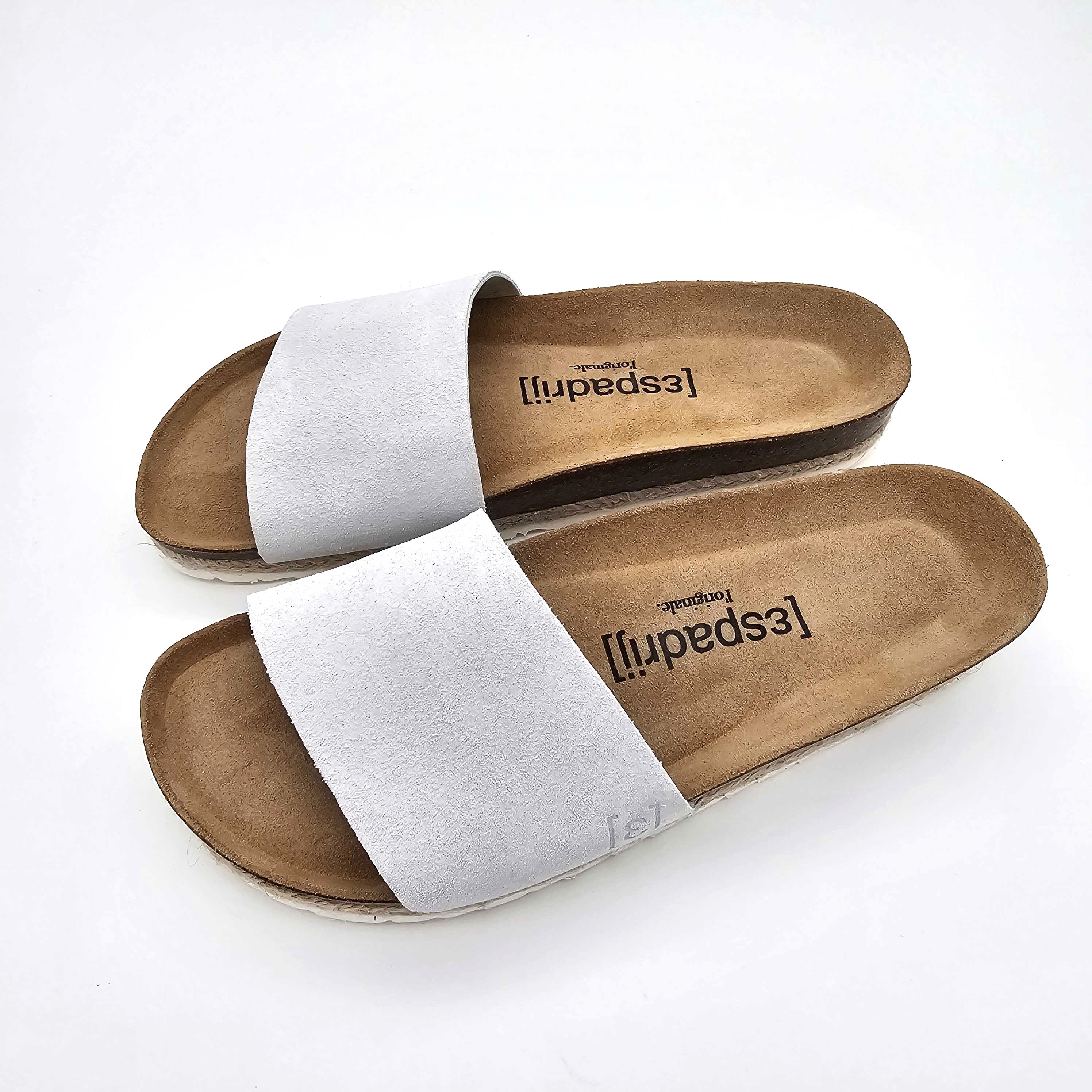 Ein Paar Schlupfsandalen mit hellbraunen Sohlen und breiten weißen Wildlederriemen auf weißem Hintergrund. Die Einlegesohlen sind mit dem Wort Espadril in schwarzer Schrift gebrandet.