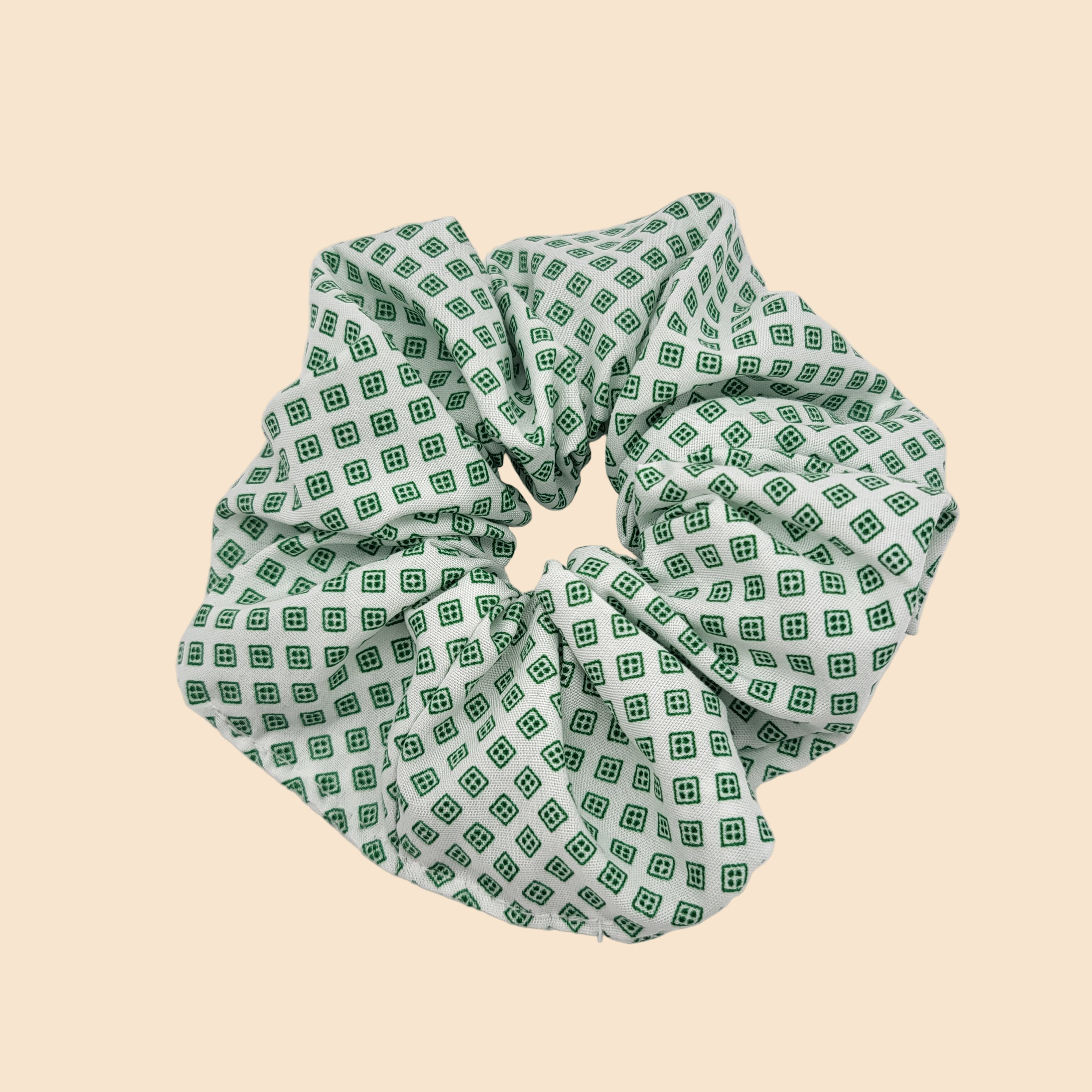 Ein Scrunchie aus Stoff mit weißem Hintergrund und grünem, geometrischem, quadratischem Muster, das auf einer schlichten weißen Fläche abgebildet ist.