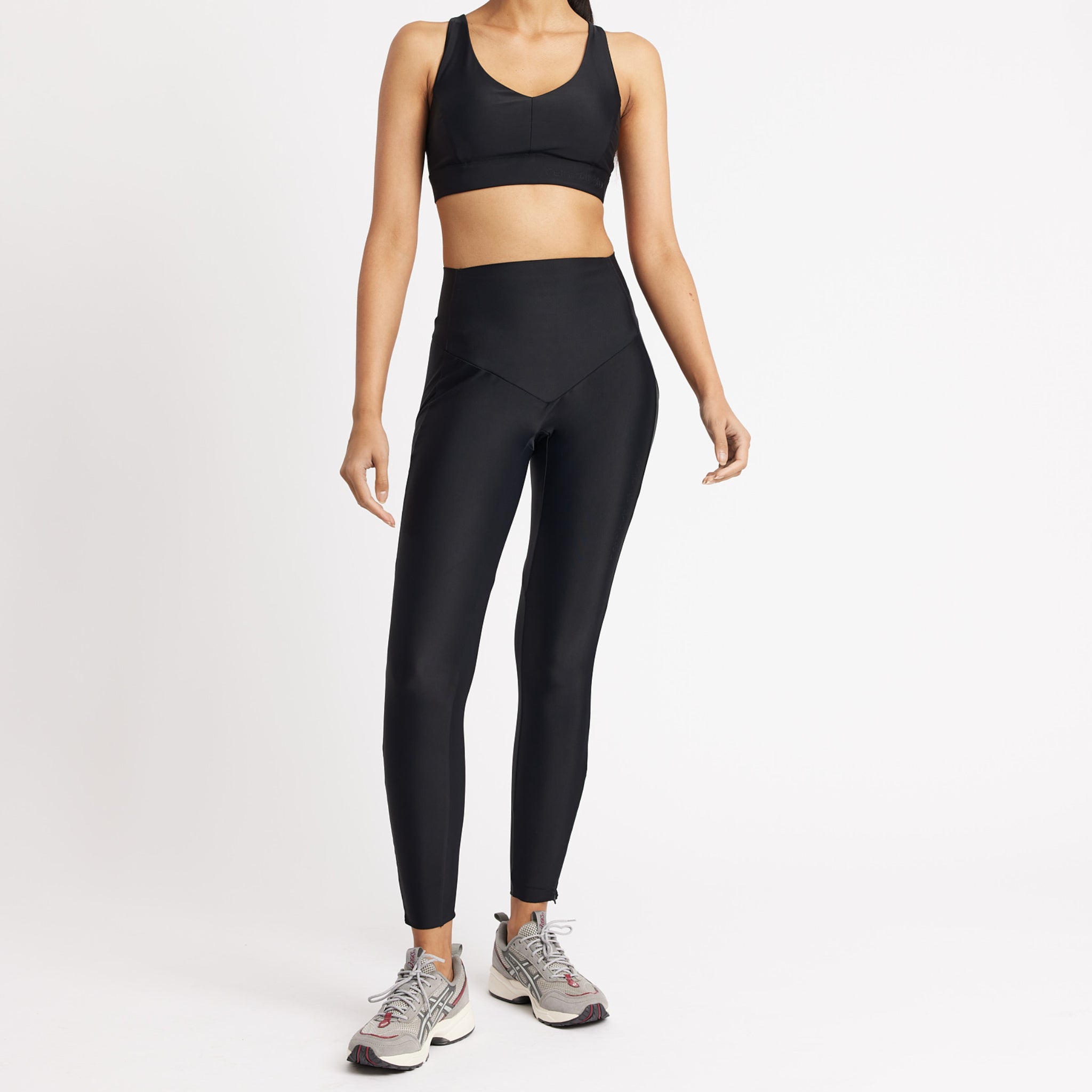 Frau in schwarzem Sport-BH und Leggings, trägt graue Sneaker vor weißem Hintergrund.