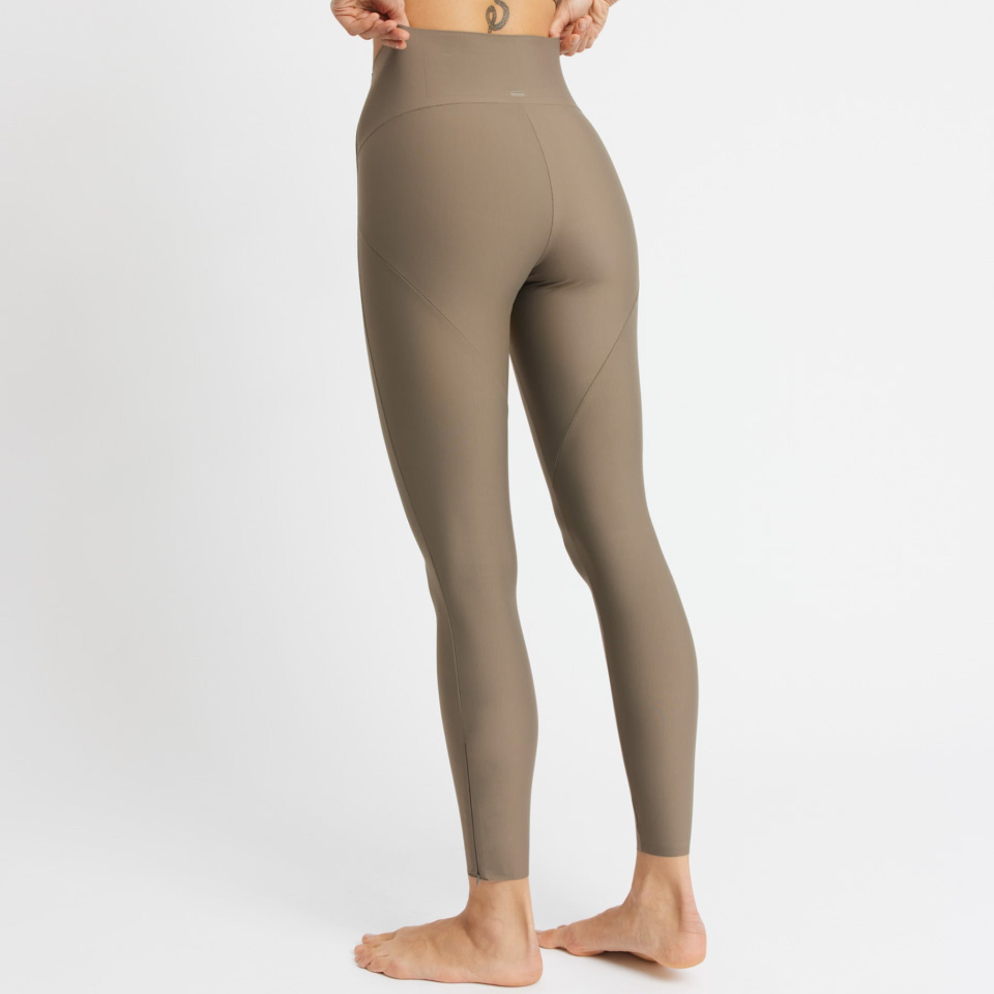 Person in taupe Leggings steht barfuß mit dem Rücken zur Kamera; ein kleines Tattoo ist über dem Hosenbund sichtbar.