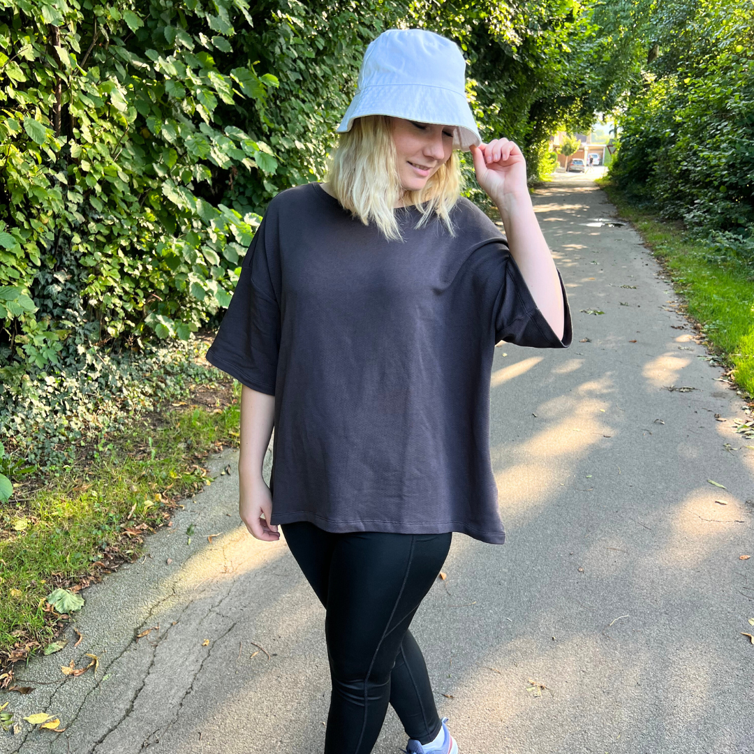 Eine Person mit weißem Hut und dunklem Shirt geht auf einem von Bäumen gesäumten Weg spazieren.