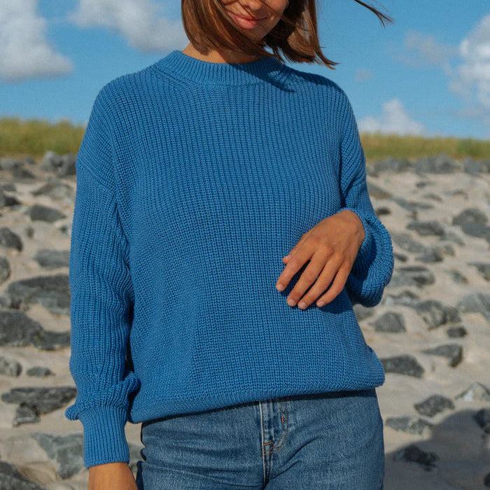 Eine Person, die einen strukturierten blauen Pullover und blaue Jeans trägt, steht im Freien auf einer felsigen Fläche mit Gras und einem teilweise bewölkten Himmel im Hintergrund. Das Gesicht der Person ist teilweise durch Haare verdeckt.