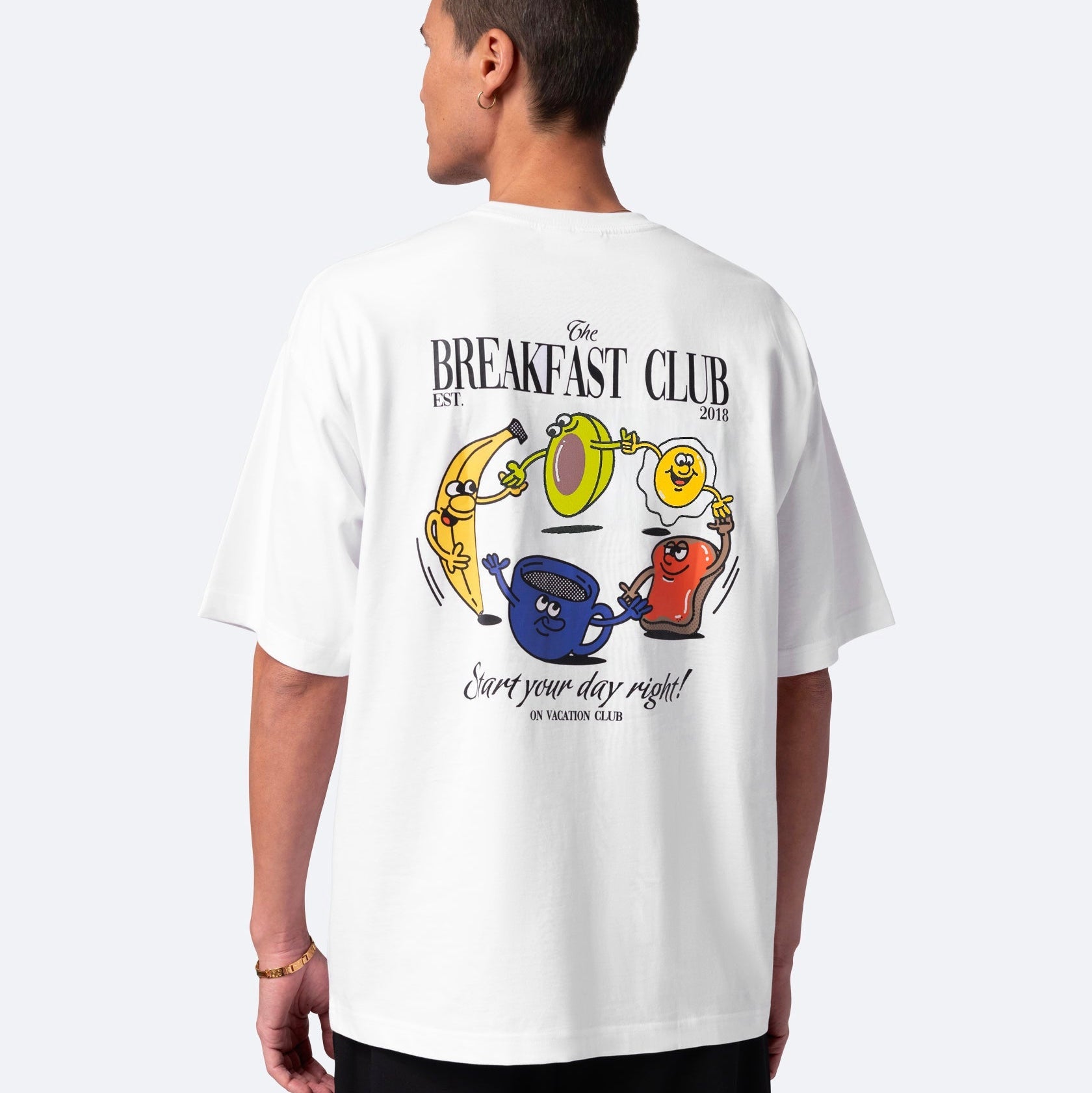 Ein Mann trägt ein weißes T-Shirt mit bunten Cartoon-Frühstücksgerichten und Tassen auf der Rückseite. Der Text lautet The Breakfast Club und Start your day right! Im Vacation Club.