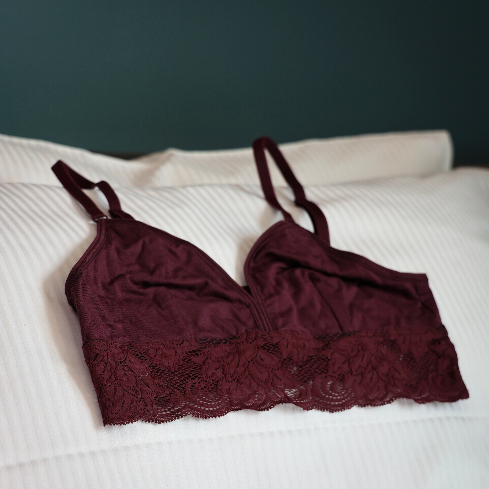 Ein bordeauxfarbener Spitzen-Bralette mit dünnen Trägern liegt auf einem weißen, gestreiften Kissen und Bettlaken.