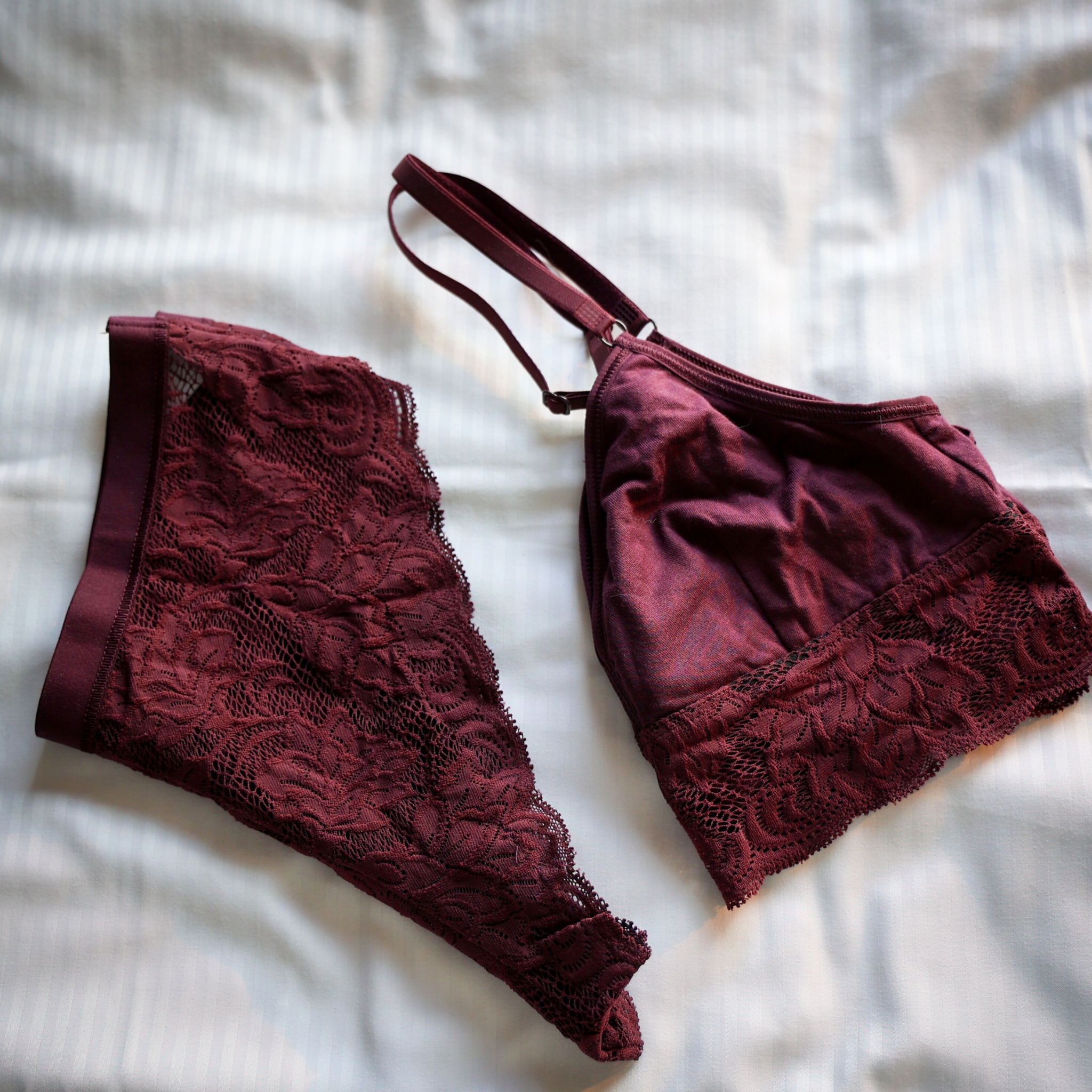 Ein passendes Set burgunderfarbener Dessous mit Spitzendetails liegt auf einem hellen, gestreiften Stoffhintergrund. Das Set besteht aus einem Bralette und einem Paar Unterwäsche.