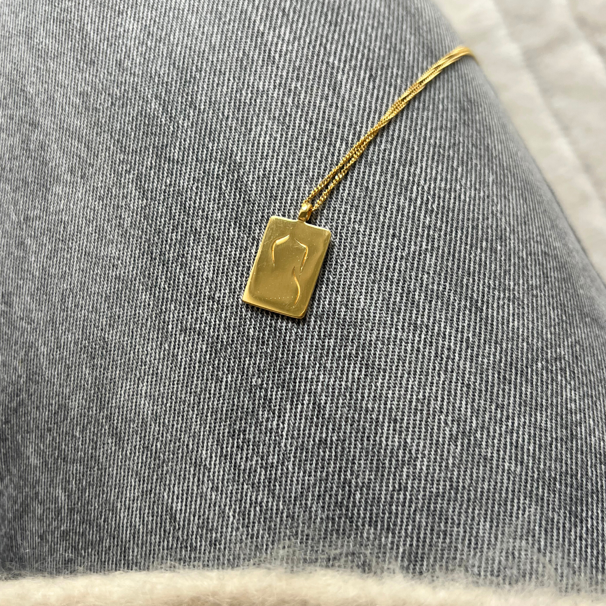 Goldene Halskette mit rechteckigem Anhänger auf grauem Jeansstoff.