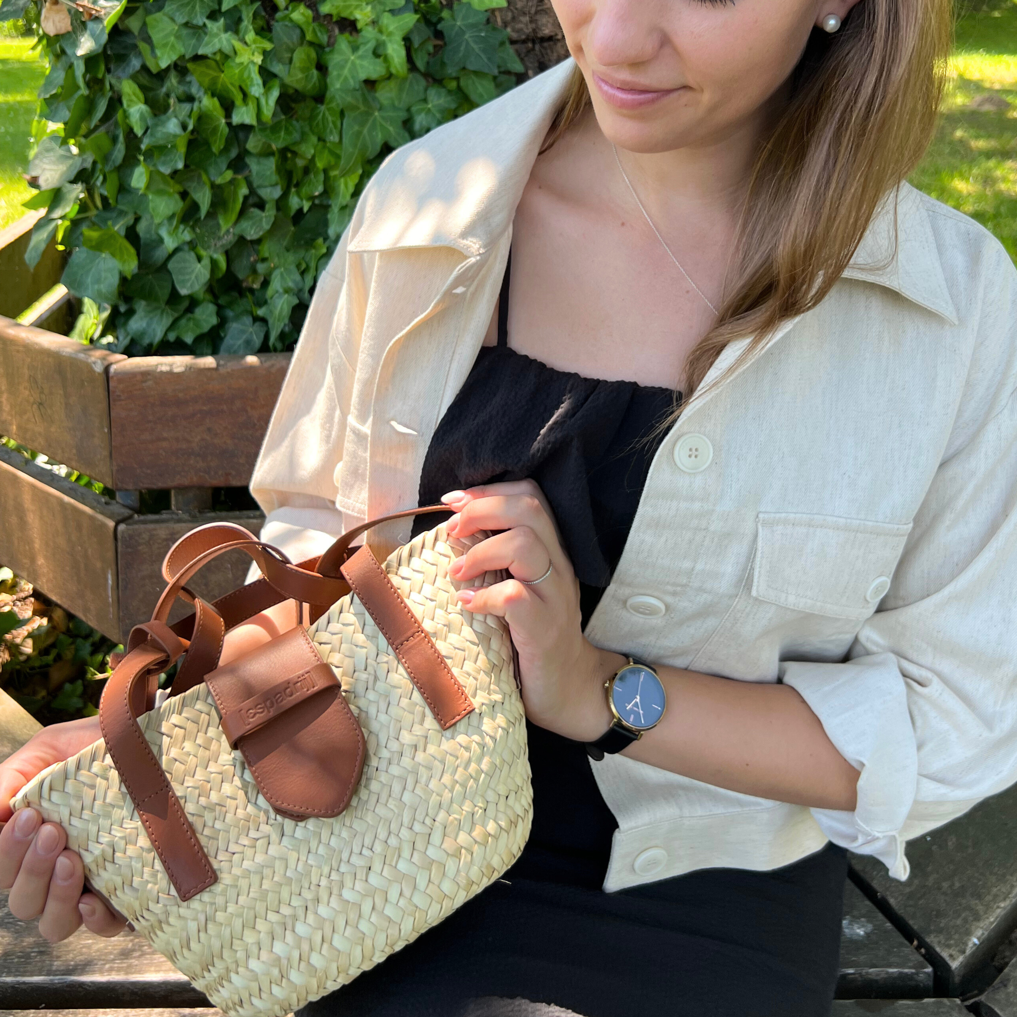 Eine Frau sitzt auf einer Holzbank im Freien und hält eine kleine geflochtene Strohtasche mit braunen Lederriemen in der Hand. Sie trägt eine cremefarbene Jacke über einem schwarzen Kleid, eine silberne Uhr und eine Halskette. Im Hintergrund ist grüner Efeu zu sehen.