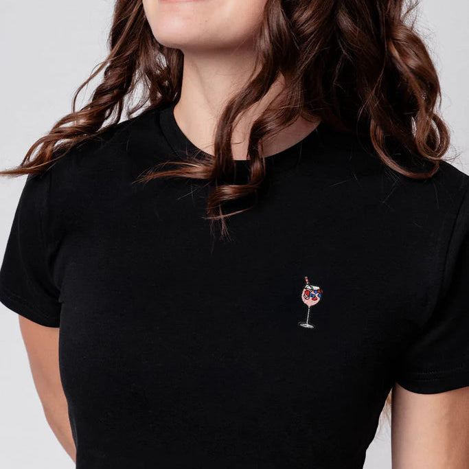 Eine Frau mit gewelltem braunem Haar und kleinen Blumenhaarspangen lächelt und blickt zur Seite. Sie trägt ein schwarzes T-Shirt mit einem aufgestickten Cocktailglas auf der Brust. Der Hintergrund ist einfarbig hellgrau.