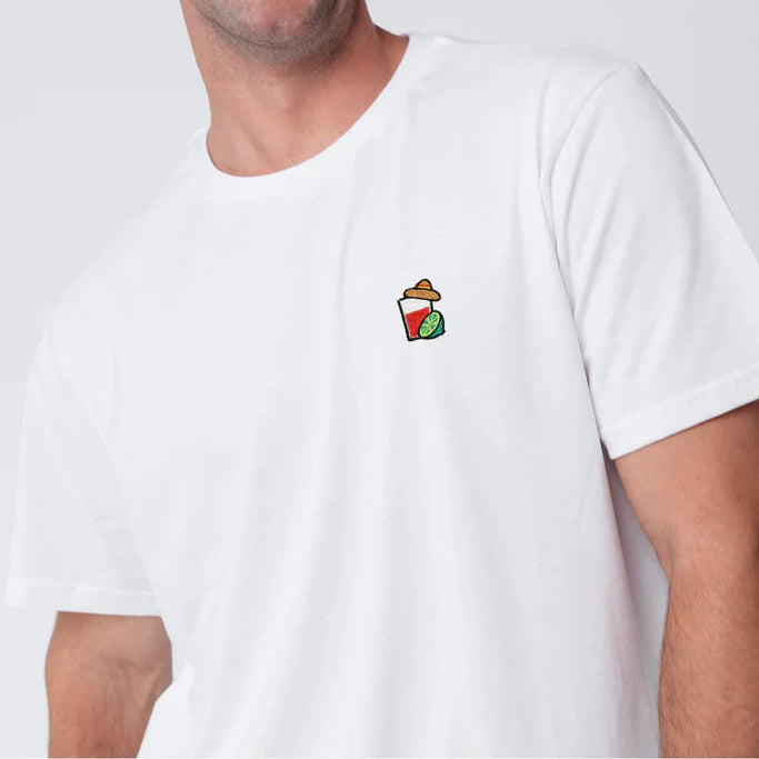 Ein lächelnder Mann mit kurzen braunen Haaren und einem Schnurrbart trägt ein schlichtes weißes T-Shirt mit einem kleinen gestickten Bild eines Getränks und einer Limette auf der Brust. Der Hintergrund ist einfarbig und in hellen Farben gehalten.
