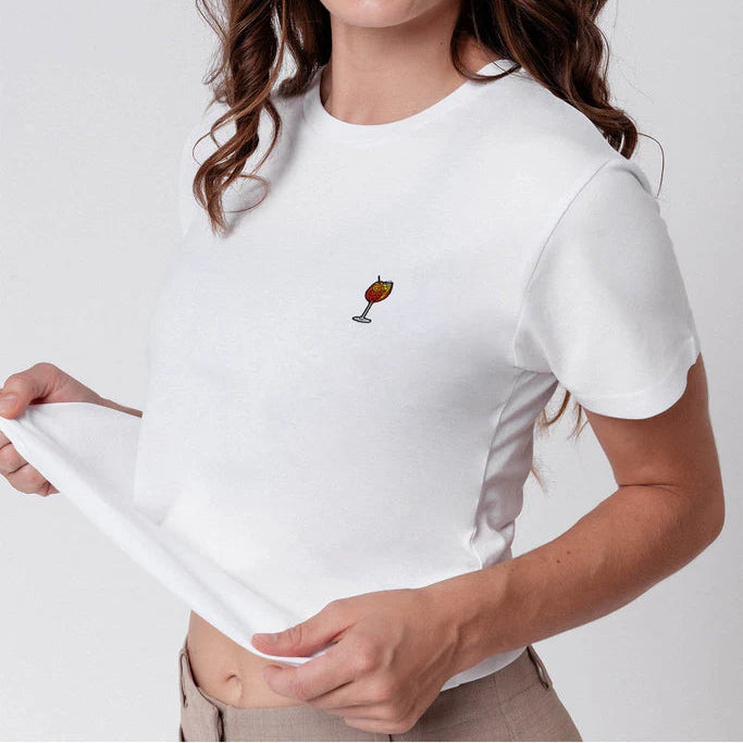 Eine junge Frau mit gewelltem braunem Haar lächelt, zwinkert und streckt die Zunge heraus. Sie trägt ein weißes T-Shirt mit einem kleinen aufgestickten Getränk und hält den Saum ihres Hemdes vor einem hellen Hintergrund.