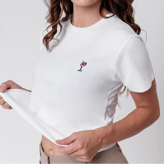 Eine Frau mit gewelltem braunem Haar und Haarspangen zwinkert und lächelt, während sie die Zunge herausstreckt. Sie trägt ein weißes T-Shirt mit einem kleinen aufgestickten Cocktailglas und hält den Saum ihres Hemdes zur Seite. Der Hintergrund ist einfarbig.